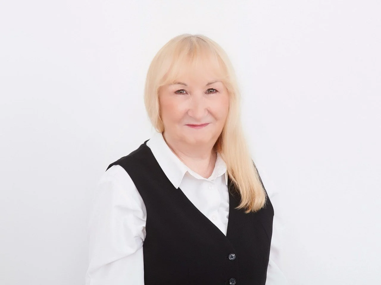 A middle-aged woman with blond hair, wearing a white shirt and black vest, smiling against a plain white background.