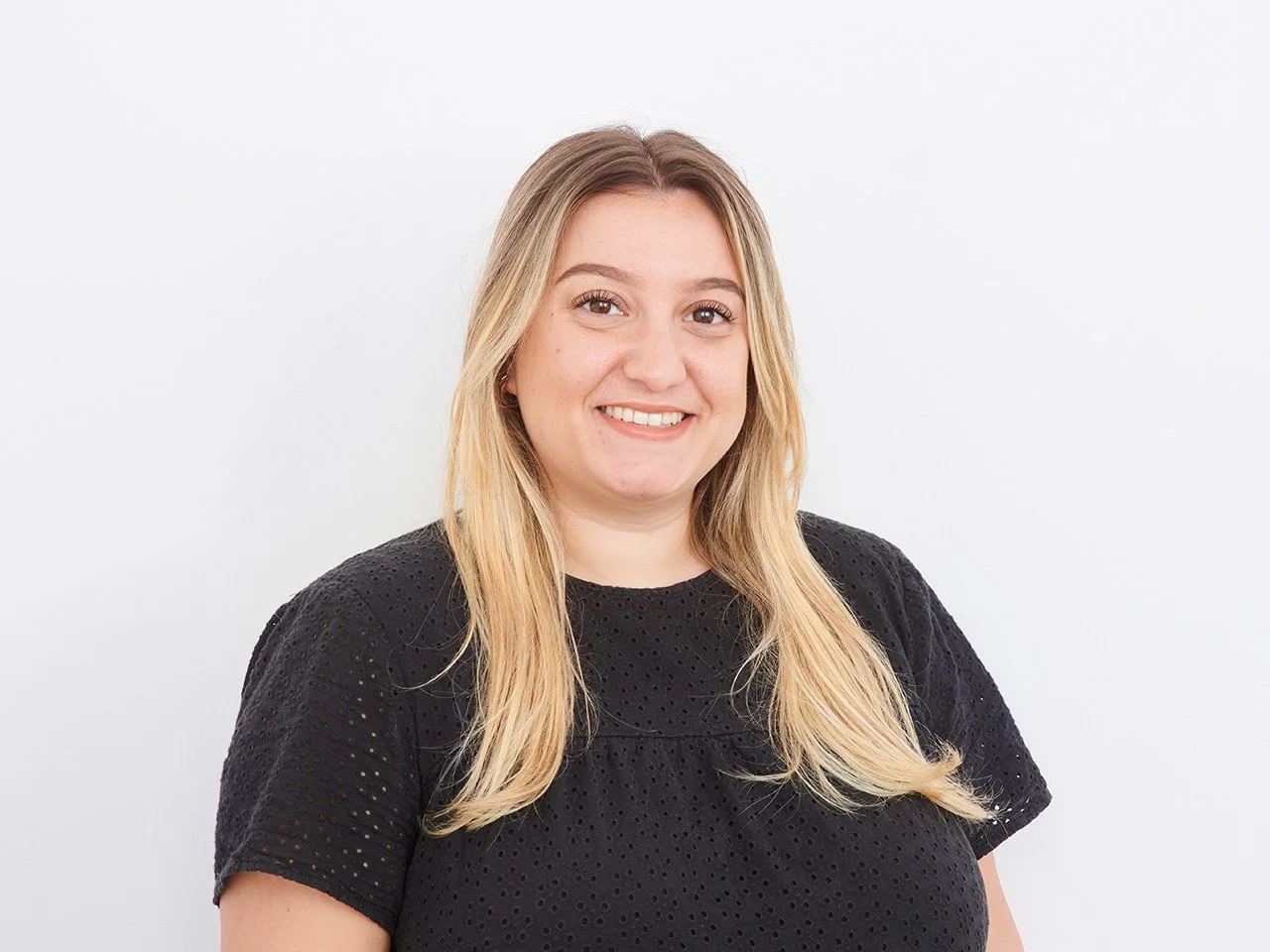 A smiling woman with long blonde hair wearing a black textured top stands against a plain white background.