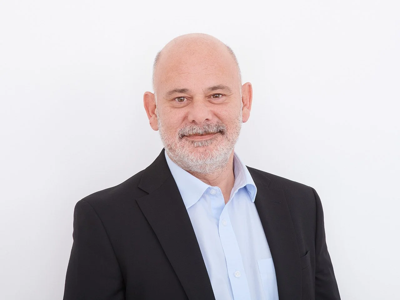 Smiling middle-aged man with light skin, a beard, and bald head, dressed in a black suit jacket and white shirt, standing against a plain white background.