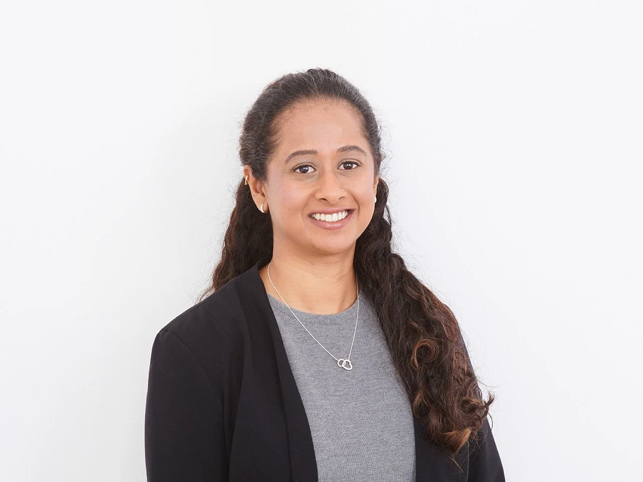 A woman with long, curly brown hair, wearing a black jacket and a silver necklace, smiling against a white background.