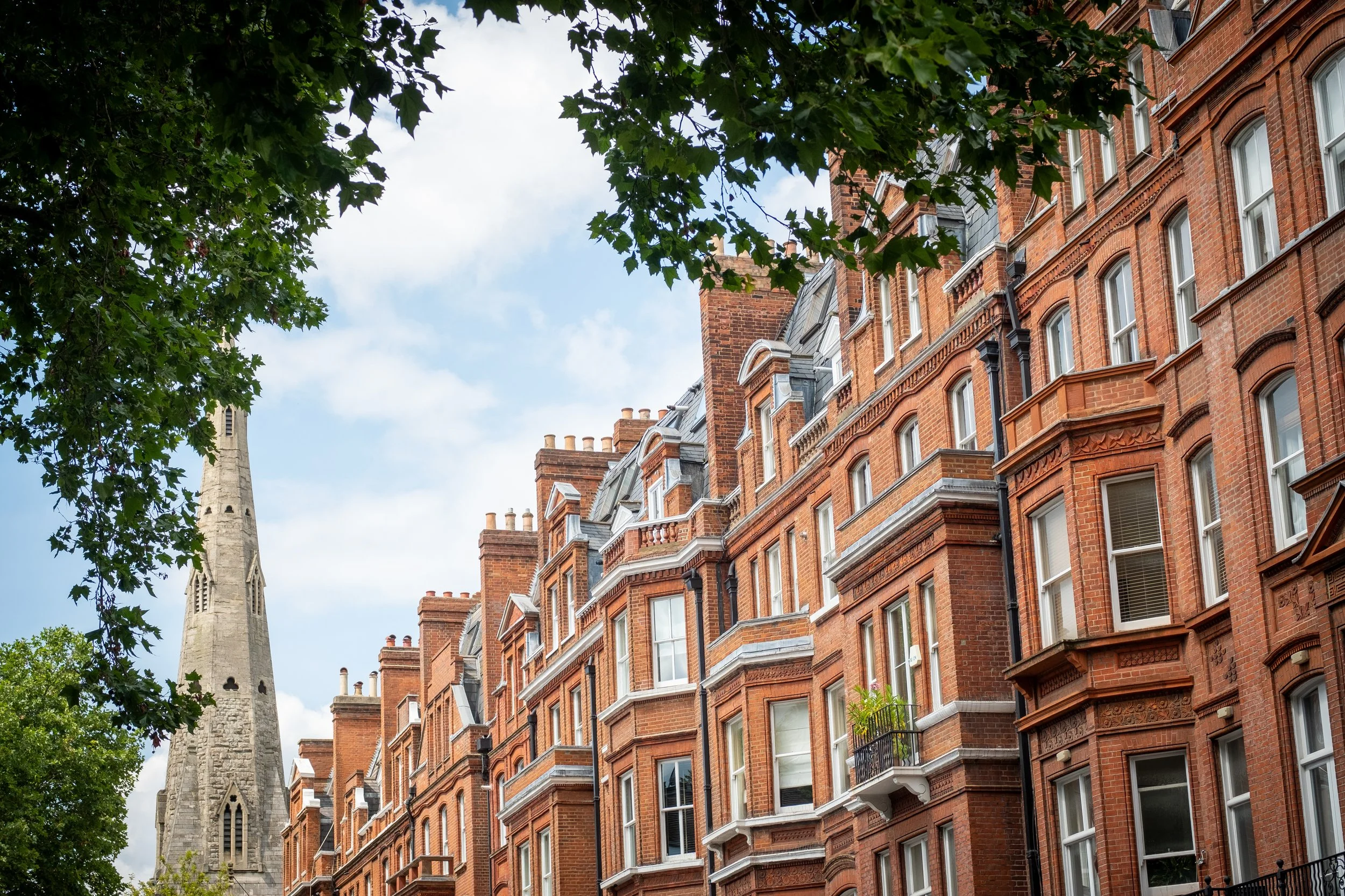 Red brick apartment buildings with bay windows, gray roofs, and a church steeple peeking from behind green trees on a partly cloudy day.