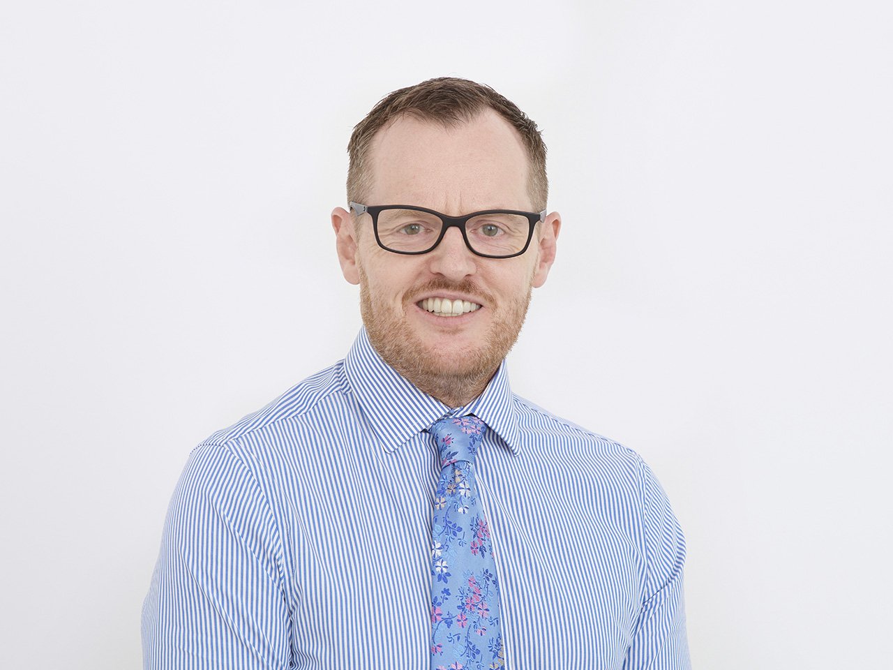 A smiling man with glasses, wearing a blue and white striped shirt and a blue floral tie, against a plain white background.