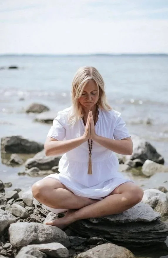 Nainen istuu rannalla, silmät suljettuina, taustalla meri ja taivas.