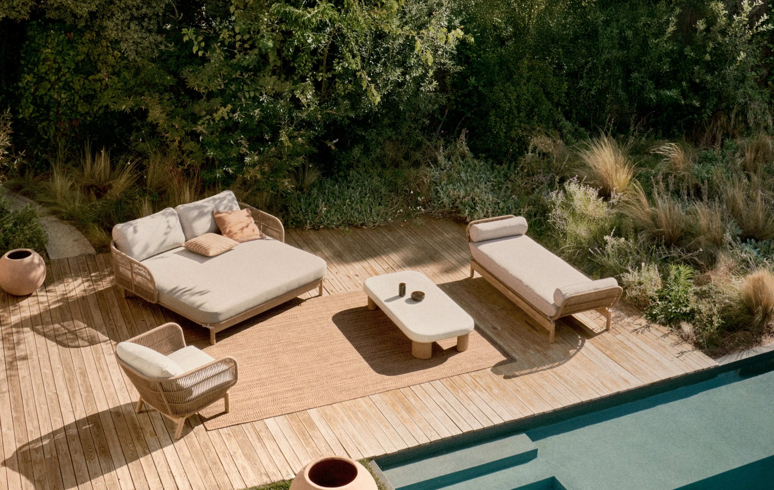 Área de lazer com móveis de varanda, incluindo sofá, cadeira, mesa de centro e espreguiçadeira, em deck de madeira ao lado de piscina, cercada por vegetação natural.