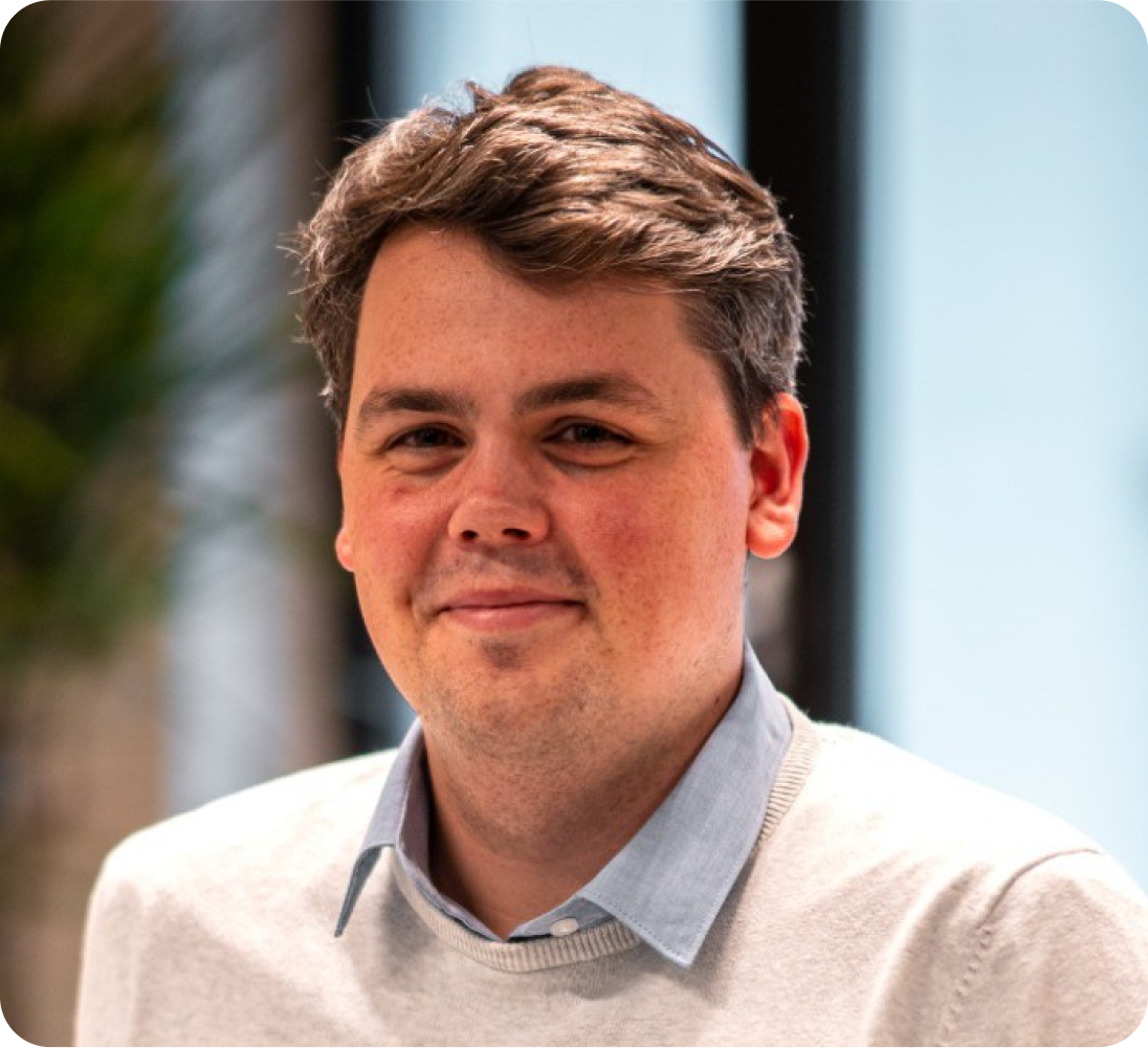 A young man with short brown hair and fair skin, wearing a light blue collared shirt under a beige sweater, smiling softly in front of a blurred indoor background.