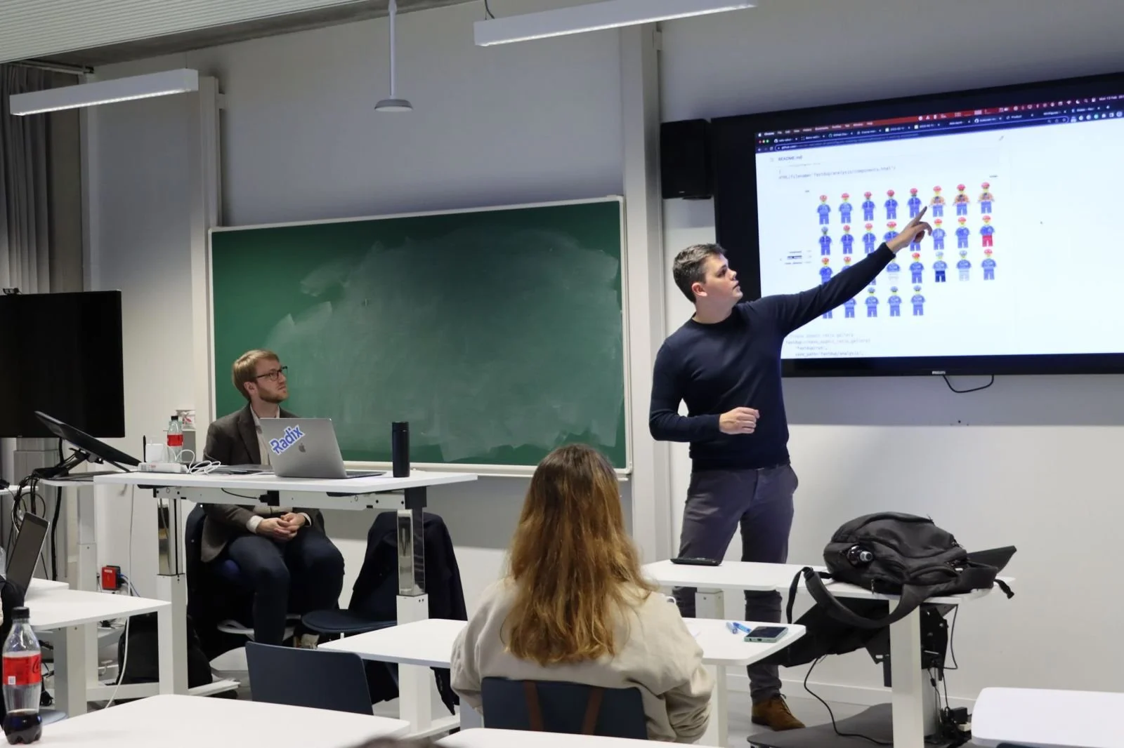 A classroom or conference room with a man giving a presentation on a large screen displaying various images of cartoon characters. Two students are visible, one sitting at a table and another with their back to the camera. A chalkboard and additional tables with laptops and bottles are also present.