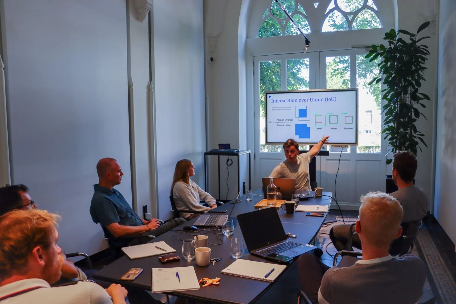 A man is giving a presentation about Venn diagrams to a group of people in a conference room with a large window and a potted plant.