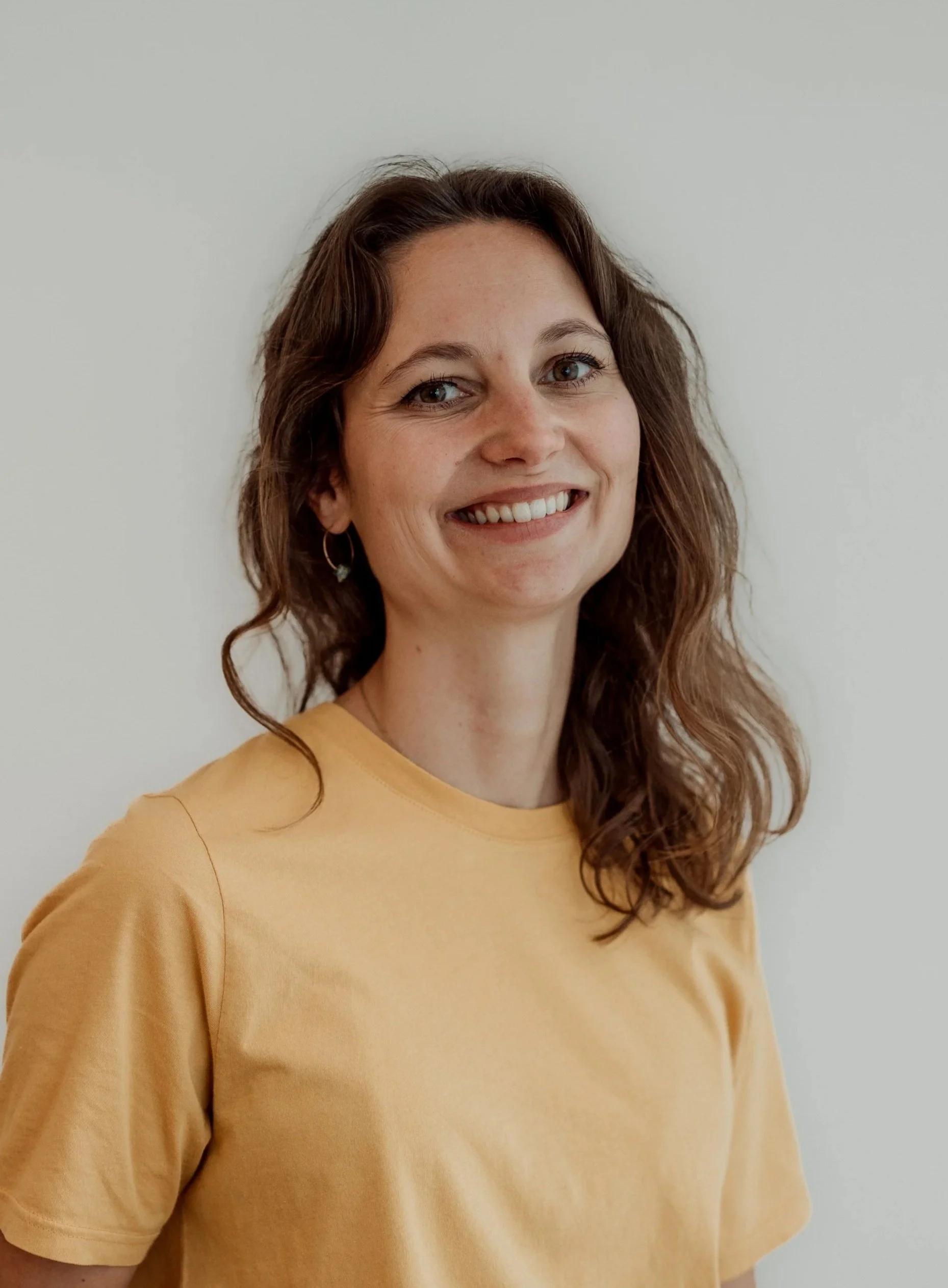 Porträt einer jungen Frau mit braunen, lockigen Haaren, blauer Augenfarbe, trägt einen gelben T-Shirt, zwei goldene Halsketten, steht vor einem Fenster in einem Raum mit unscharfem Hintergrund.