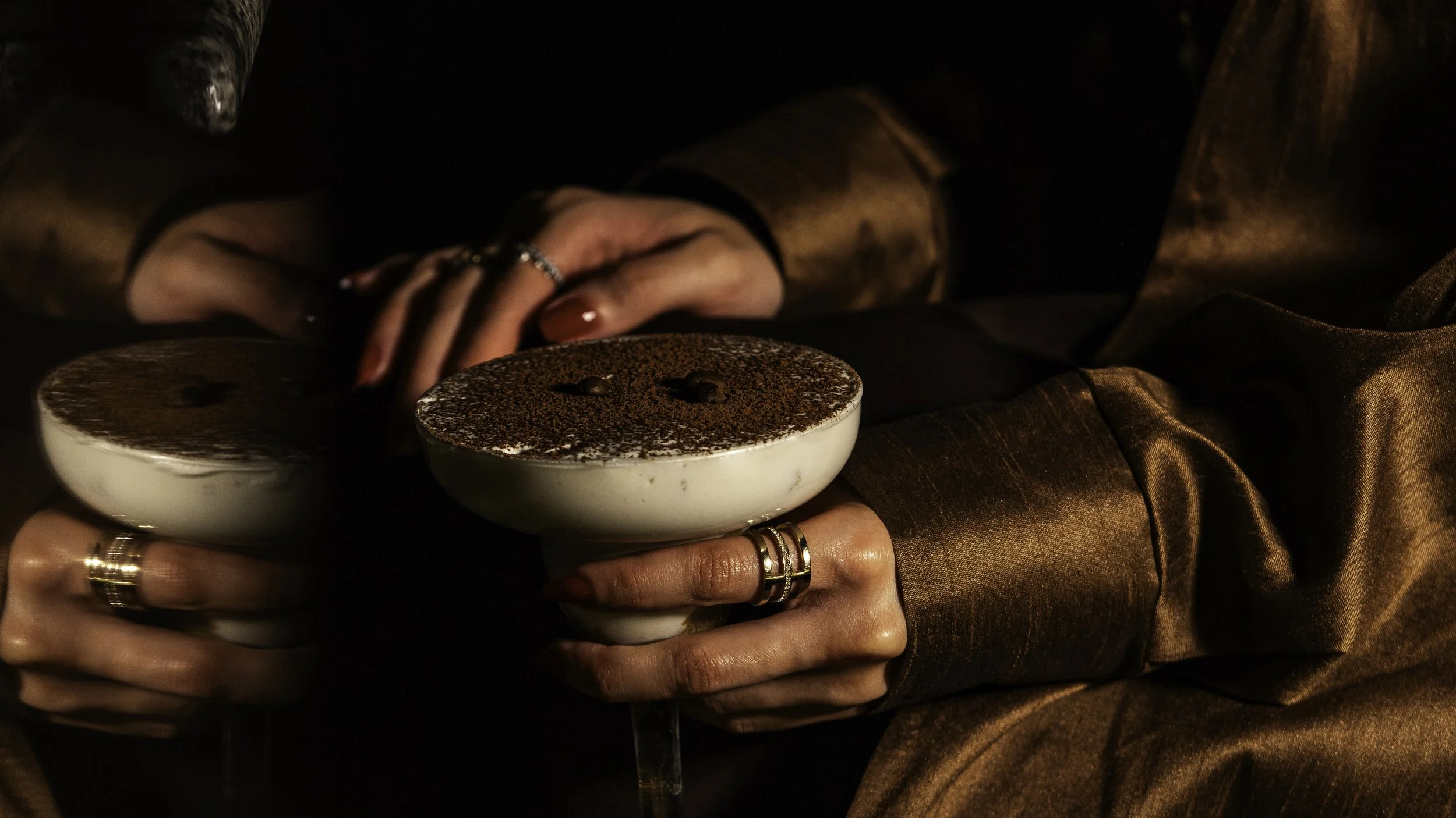 Two women holding white cups of tiramisu topped with cocoa powder, in a dimly lit setting.