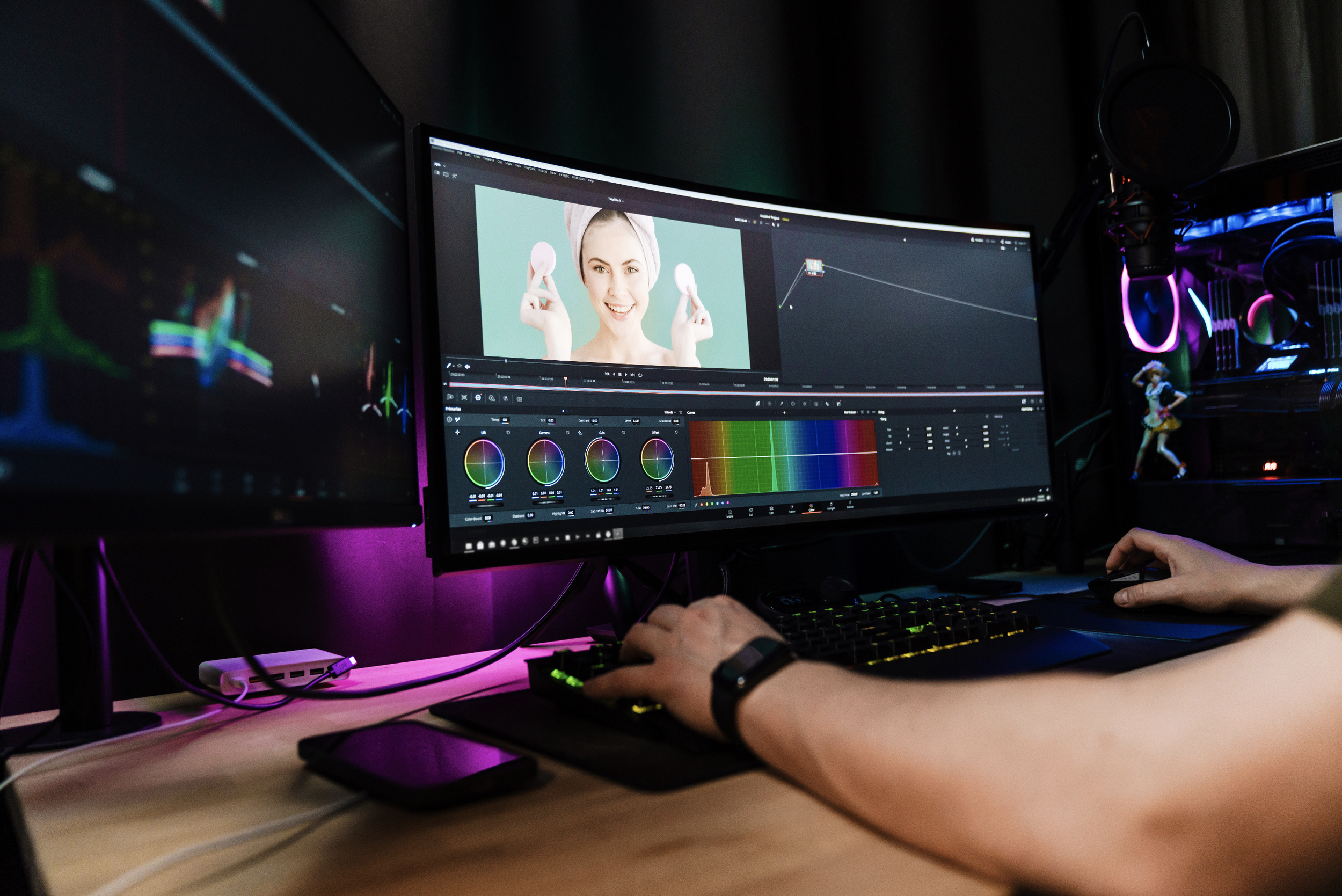Person working on video editing in a dark room with multiple monitors, colorful RGB lighting, and editing software on the screen.