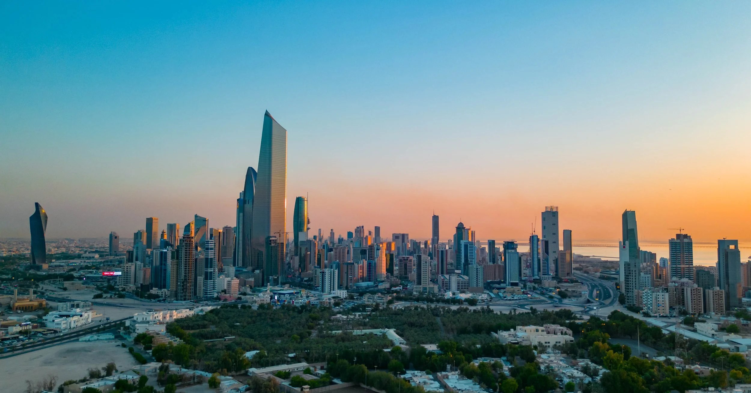 A city skyline at sunset with numerous tall skyscrapers, including a prominent pointed building, and a river in the background.
