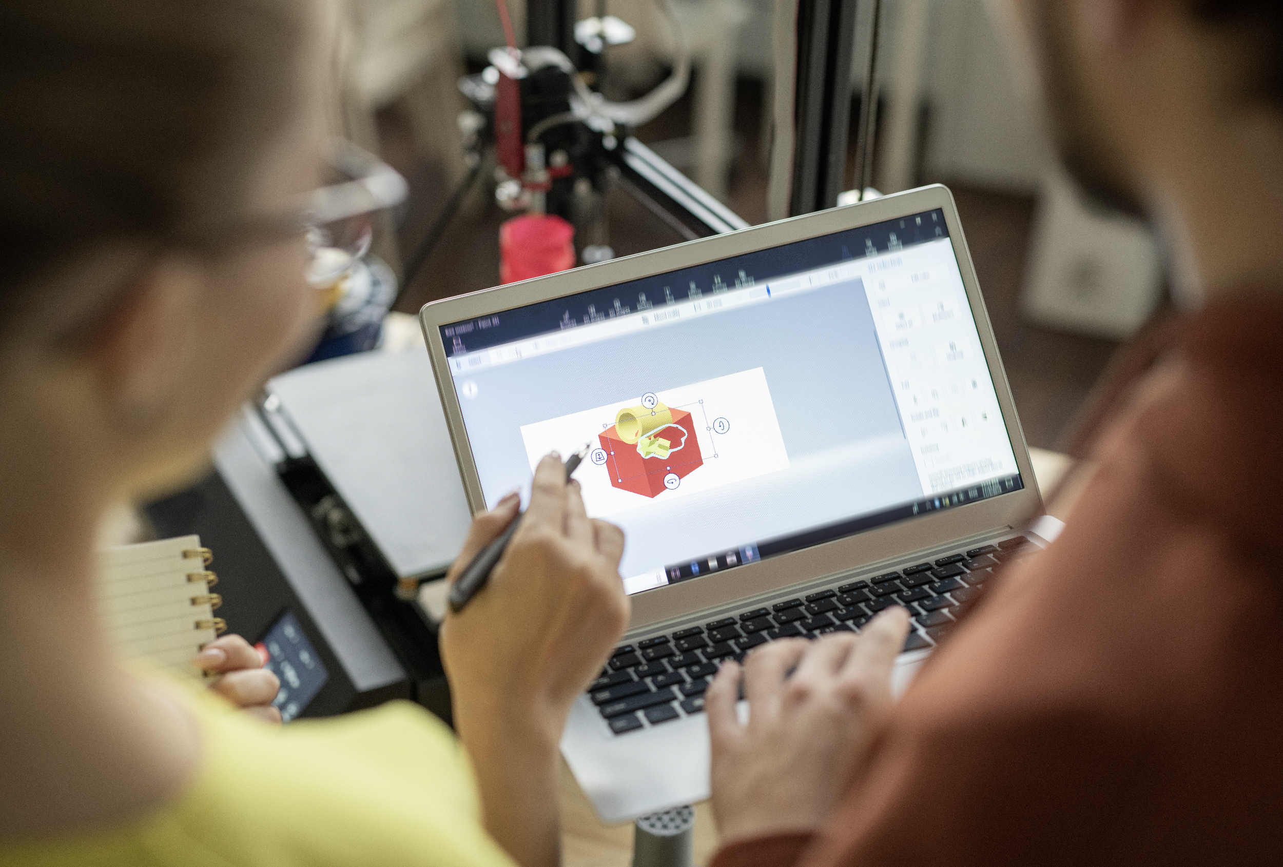 Two people observe a laptop screen displaying a 3D design of a red cube with yellow and green objects on it, in a workshop setting.