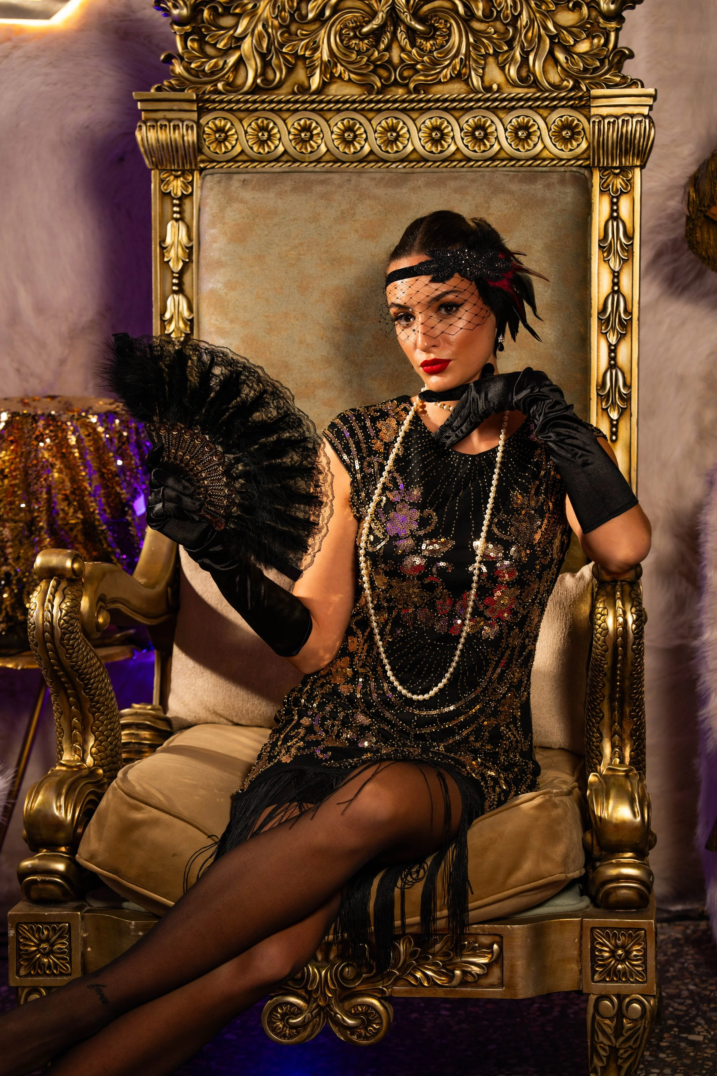 A man and a woman in vintage costumes seated on ornate gold chairs in a luxurious room with eclectic decor, neon sign, and artful lighting.