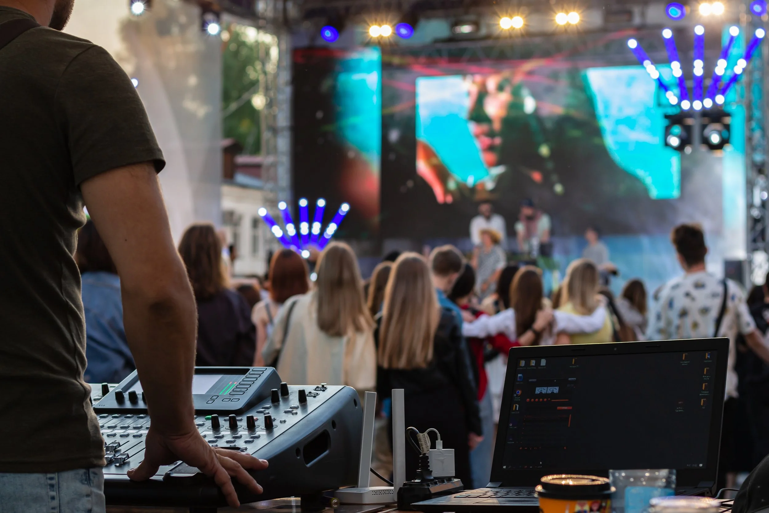 A DJ controlling sound equipment at an outdoor concert with a large crowd watching a stage featuring performers and colorful screens.