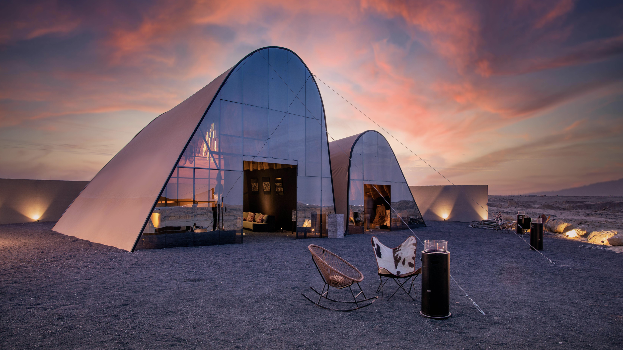 Modern glass-domed structures with curved roofs set in a desert landscape at sunset, with outdoor seating and lighting.