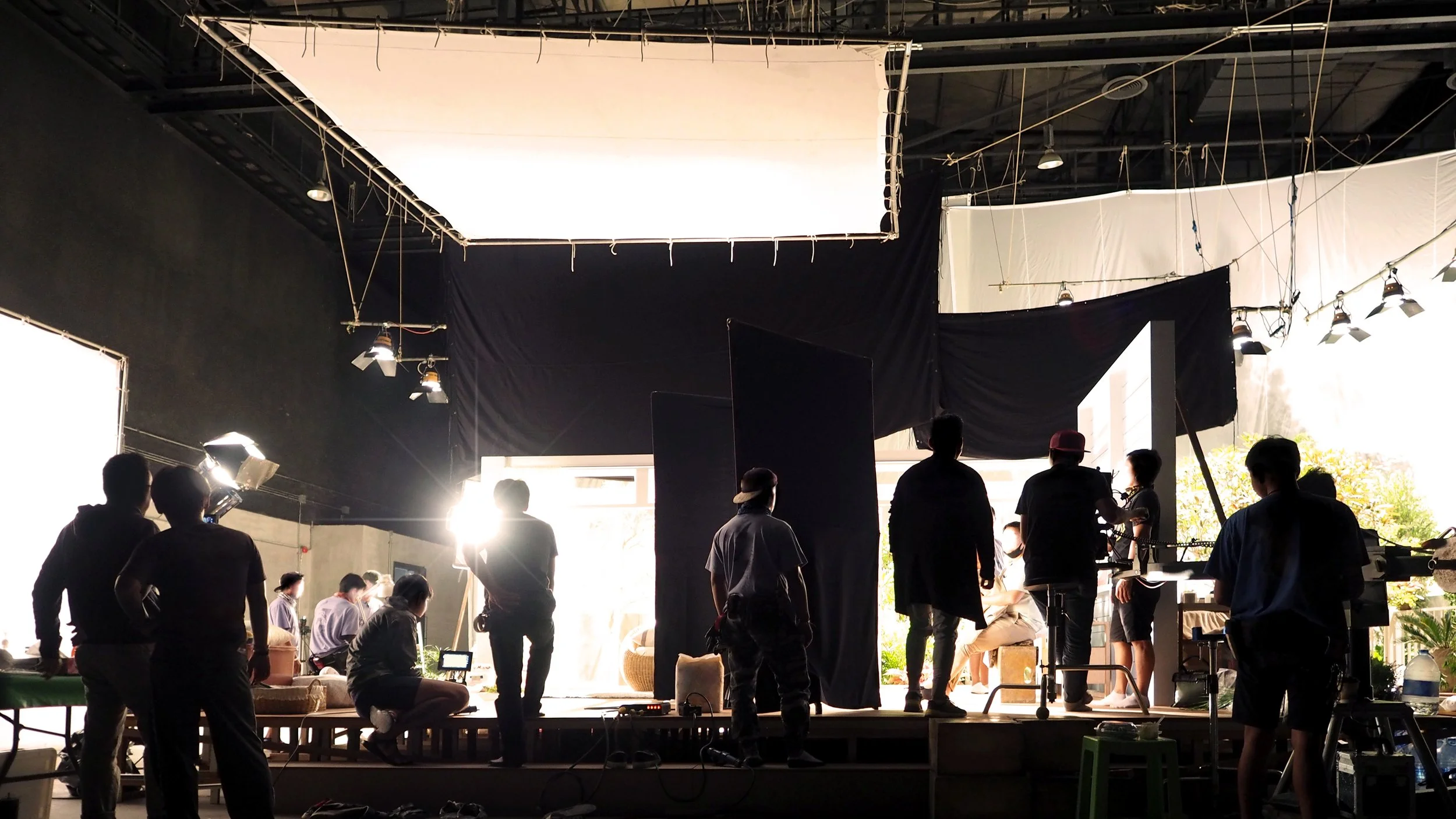 Film or photo shoot set with crew members, camera equipment, lighting, black curtains, and large windows letting in natural light.