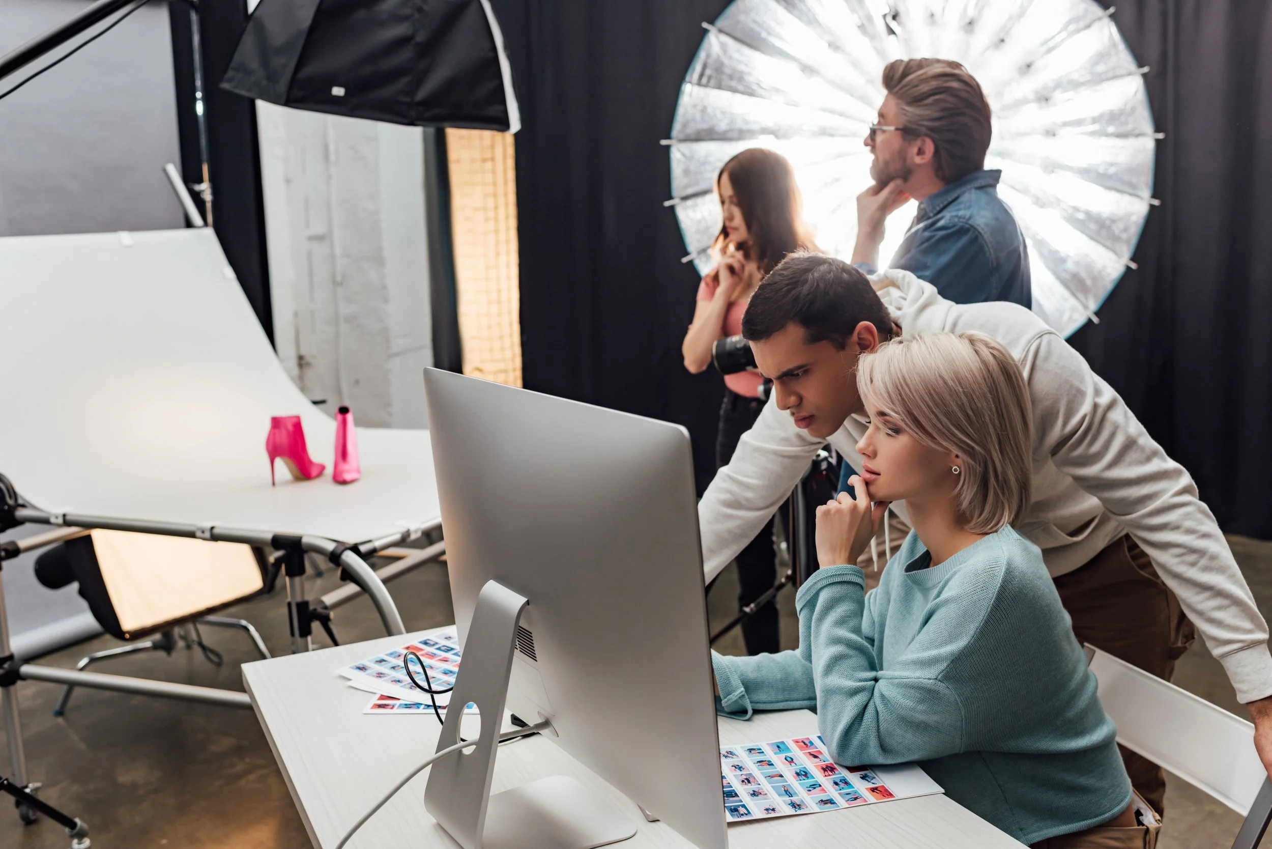 Photography studio with four people working on a photo shoot. A woman in a light blue sweater is seated at a computer, while a man in a white hoodie leans over her shoulder. In the background, a woman with long hair and a man with sunglasses are standing in front of a large silver umbrella reflector, with professional lighting and a white backdrop visible.