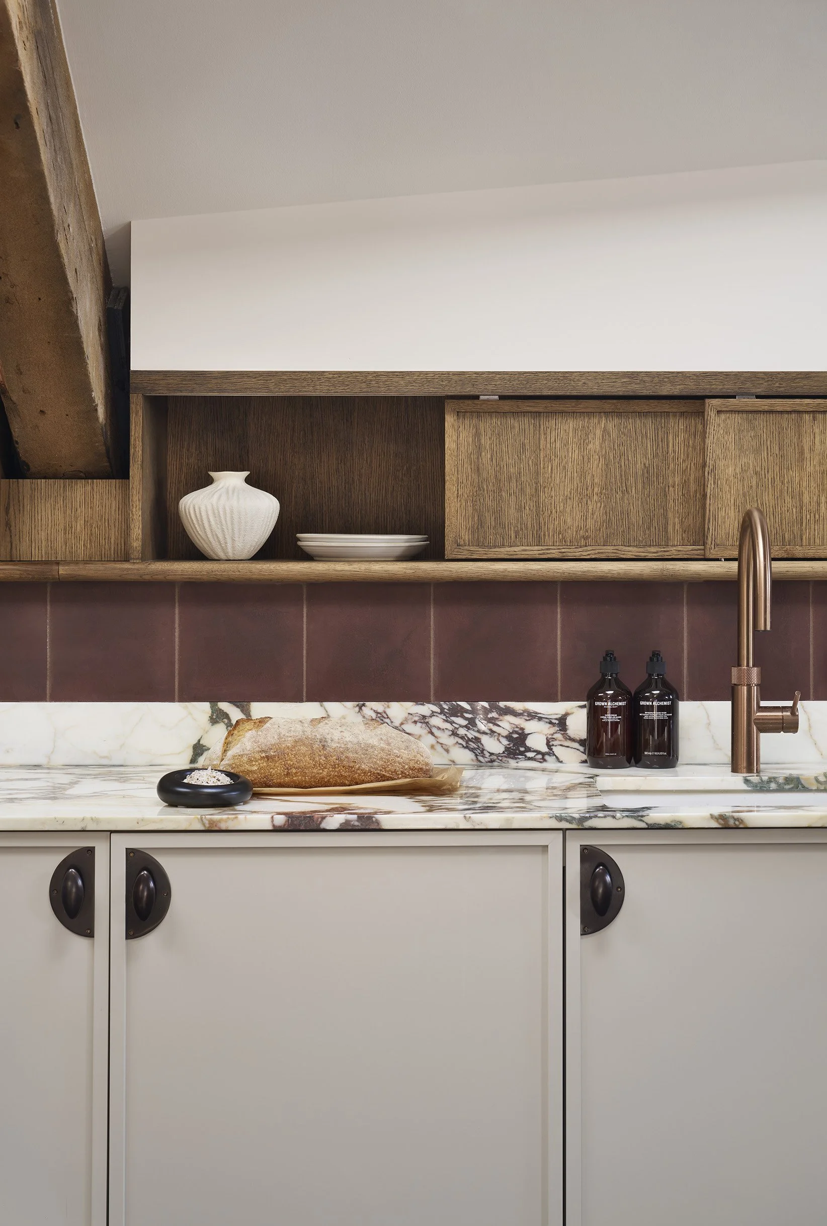 A kitchen countertop with a loaf of bread, a black dish with salt, two amber bottles with black caps, a bronze faucet, and wooden cabinetry with decorative plates and a vase.