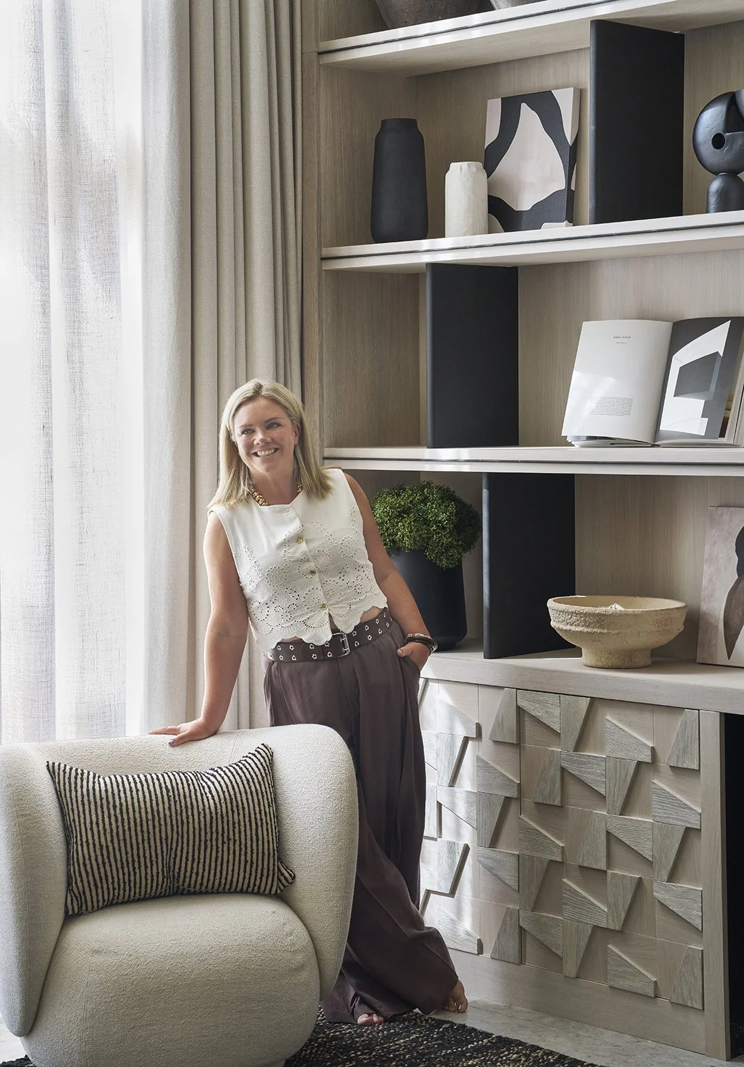 A woman standing in a modern living room next to a cream-colored armchair with a striped pillow, in front of a built-in shelf with decorative objects and books, with beige curtains and natural light.