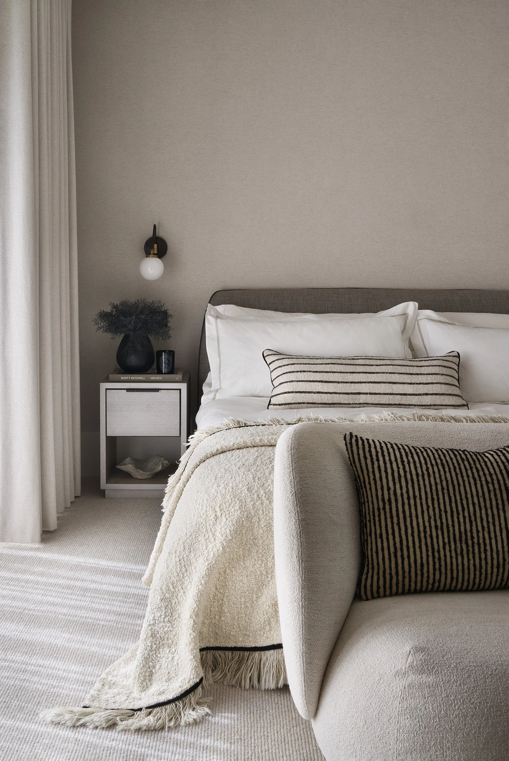 A styled bedroom corner with a bed, beige bedding, a black and white striped throw pillow, a side table with a black vase containing greenery, a small black object, and a shelf with books and decor. There is a wall-mounted light fixture and beige curtains on the left.