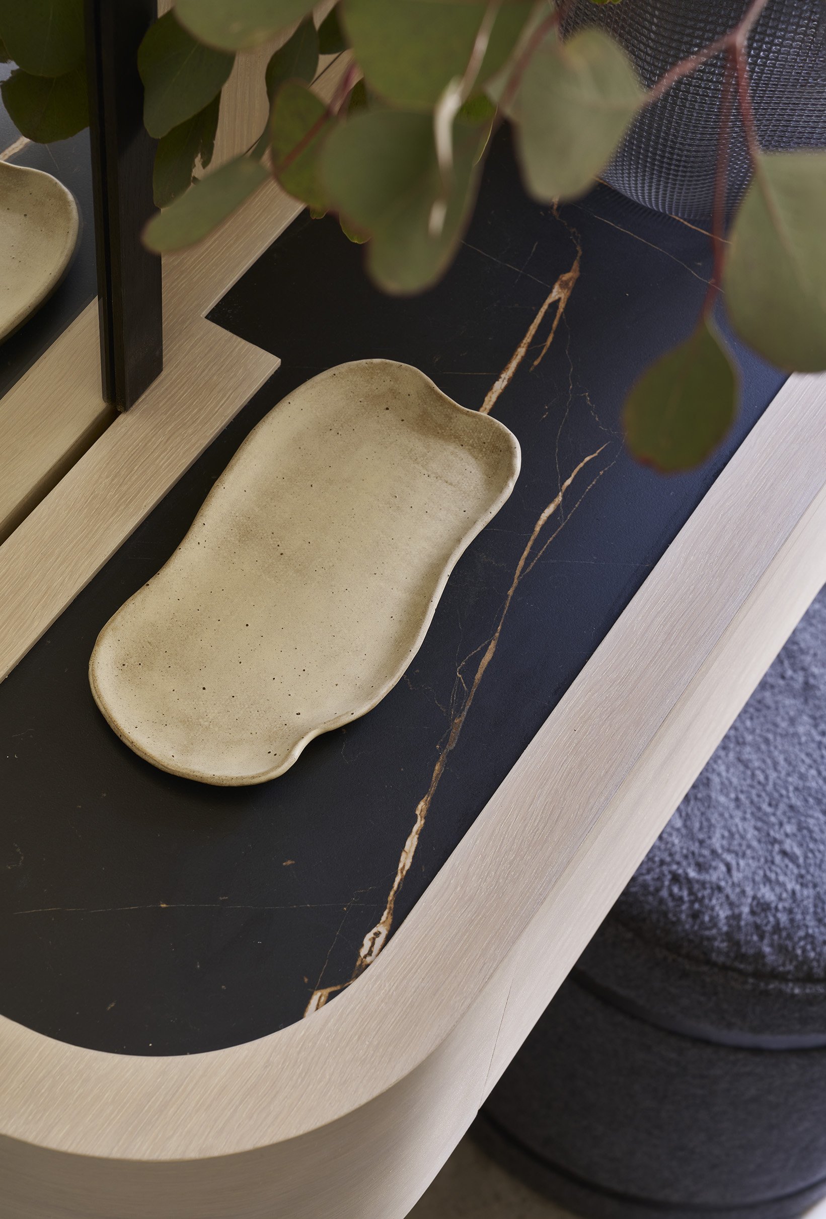 A rectangular, beige ceramic tray with rounded edges on a black marble table with gold veining, next to a green leafy plant and a mirror reflecting part of the scene.