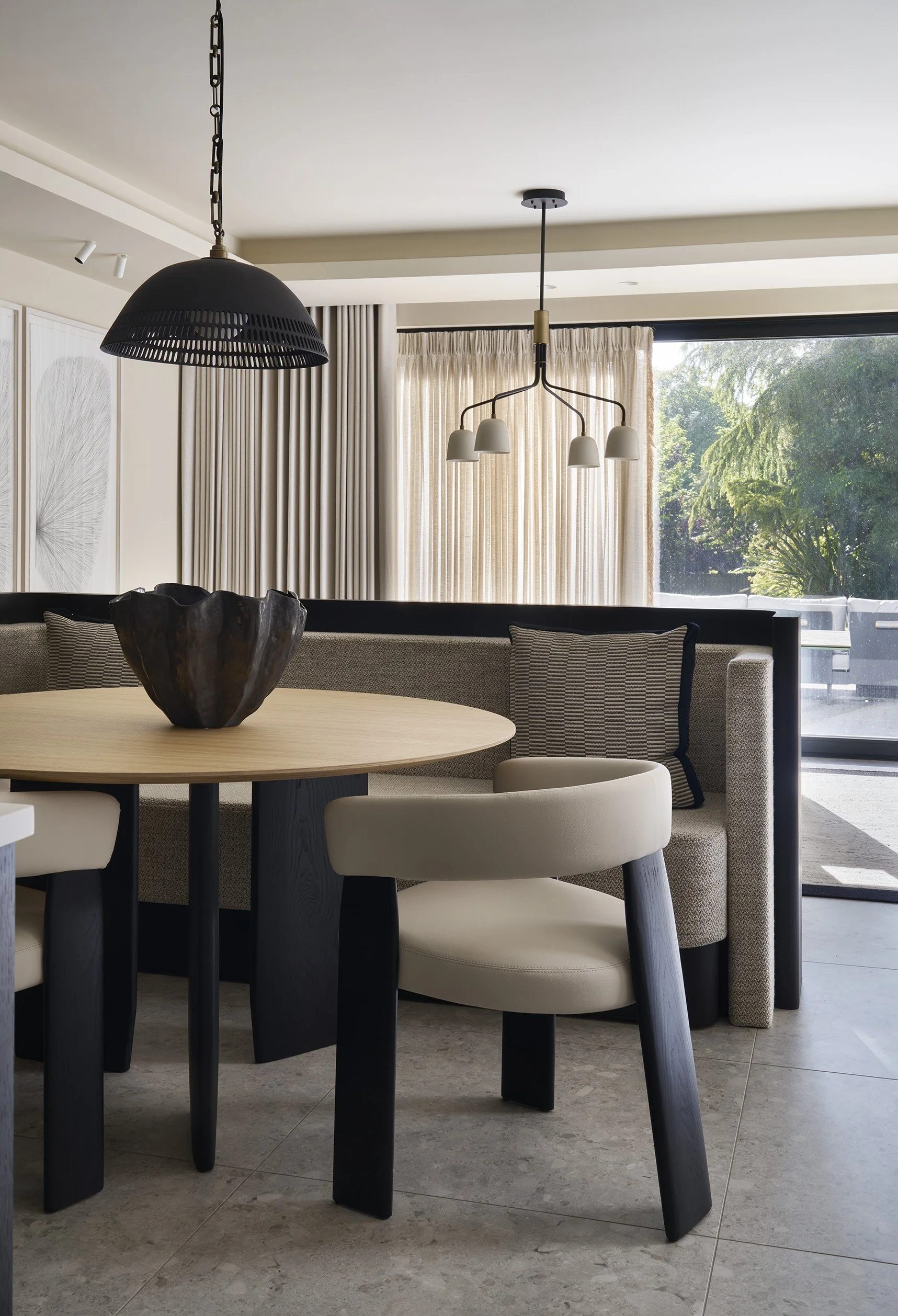 Modern dining area with a round wooden table, beige chairs, a black pendant light, decorative bowl, and a built-in cushioned bench with striped and plain pillows, next to a window with beige curtains.