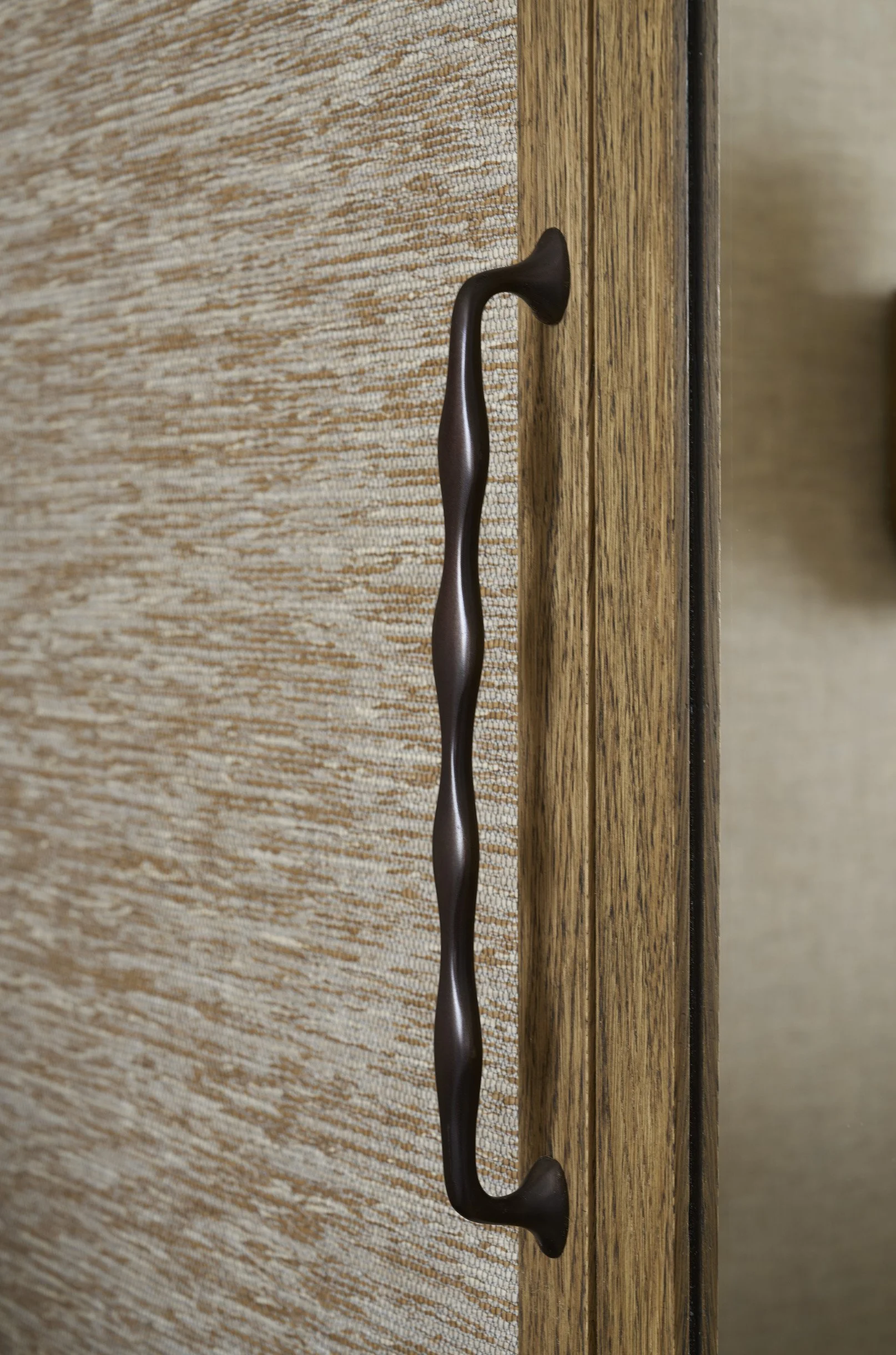 Close-up of a dark brown, wavy cabinet or drawer handle mounted on a wooden surface with a textured fabric or wallpaper in the background.