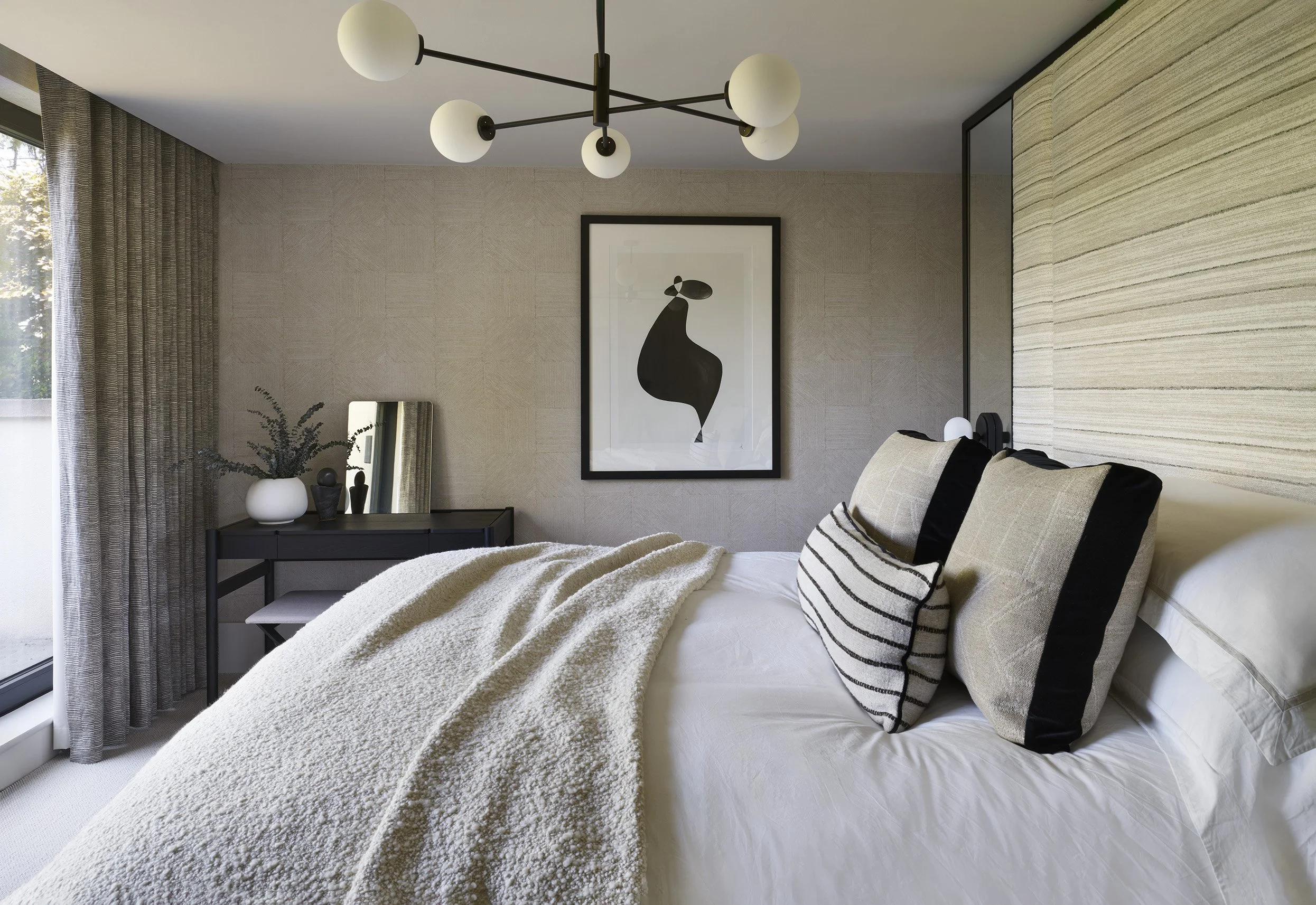 Modern bedroom with a bed, striped pillows, beige textured wall, black and white abstract artwork, black dresser with a mirror, potted plant, gray curtains, and a ceiling light fixture with globe bulbs.