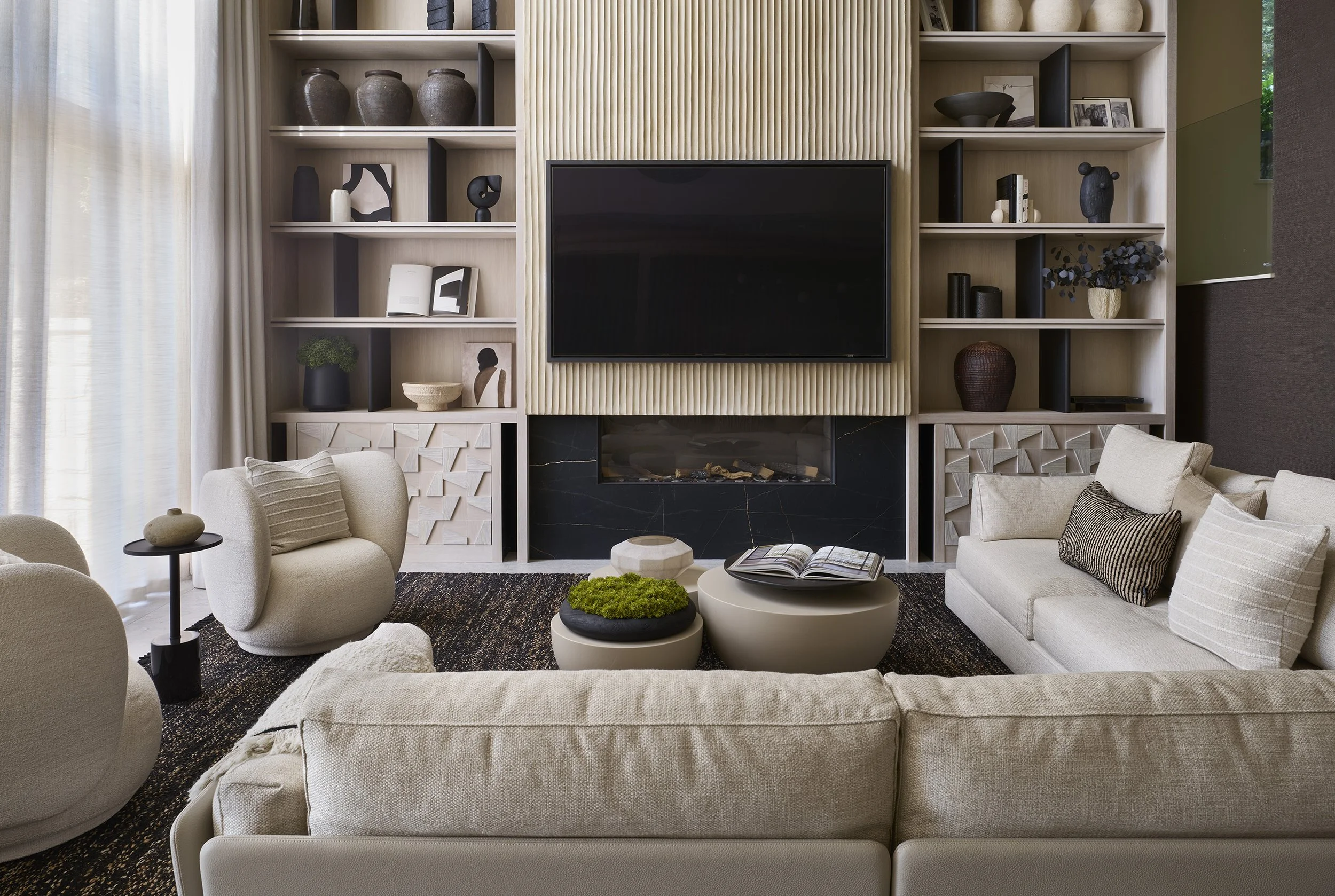 Modern living room with beige sofa and chairs, black and beige coffee tables, a wall-mounted TV, and built-in shelves with decorative items.