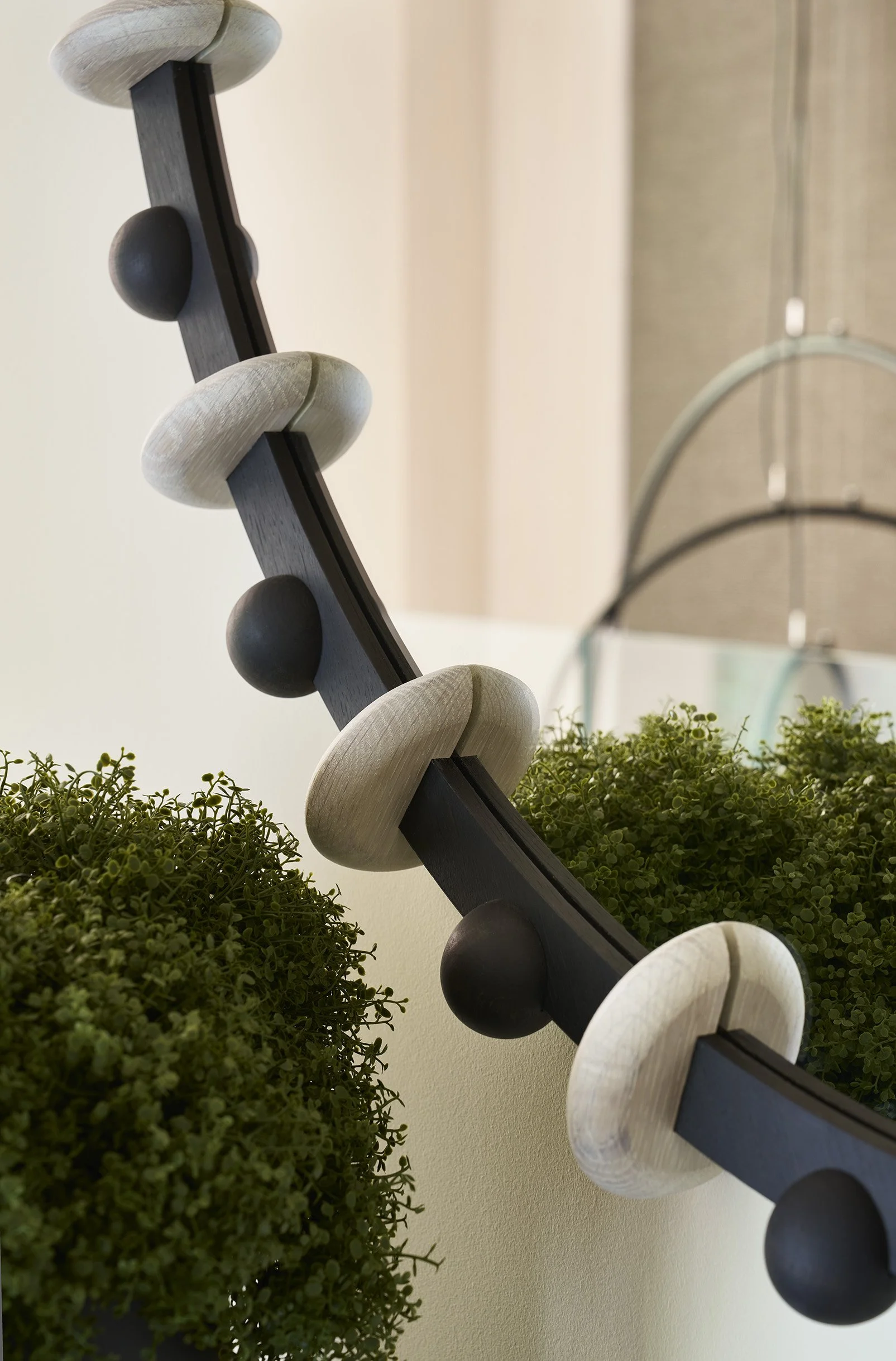 Decorative wall art with a curved black piece adorned with alternating white wood and dark spherical accents, surrounded by green plants and mirror reflections.