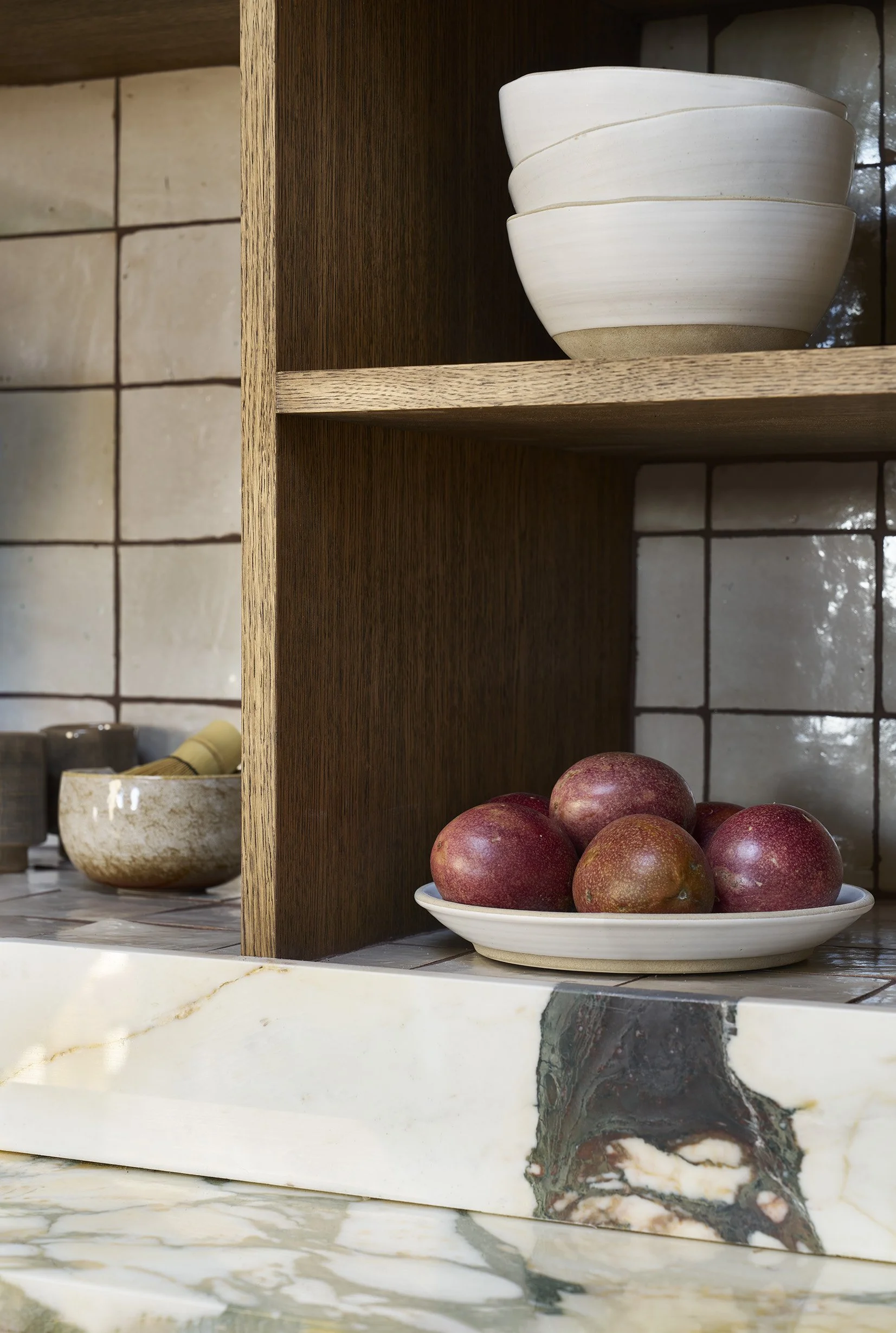 A kitchen shelf with stacked white bowls and a white plate with apples on a marble countertop against a tiled wall.