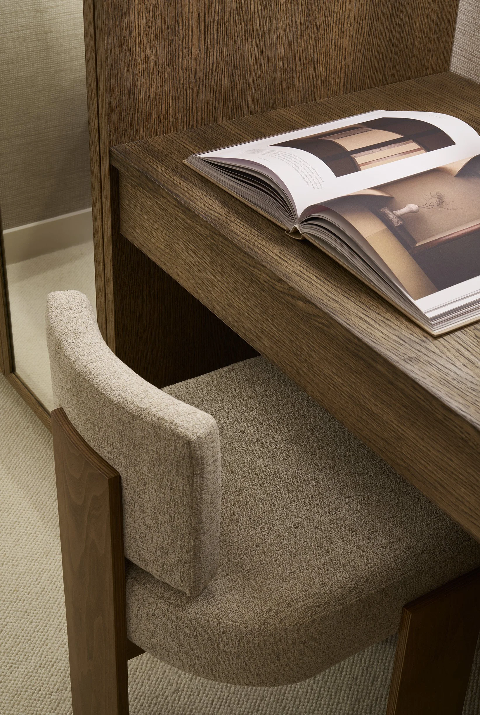 Open book on a wooden desk, with a beige upholstered chair and a glass partition behind.
