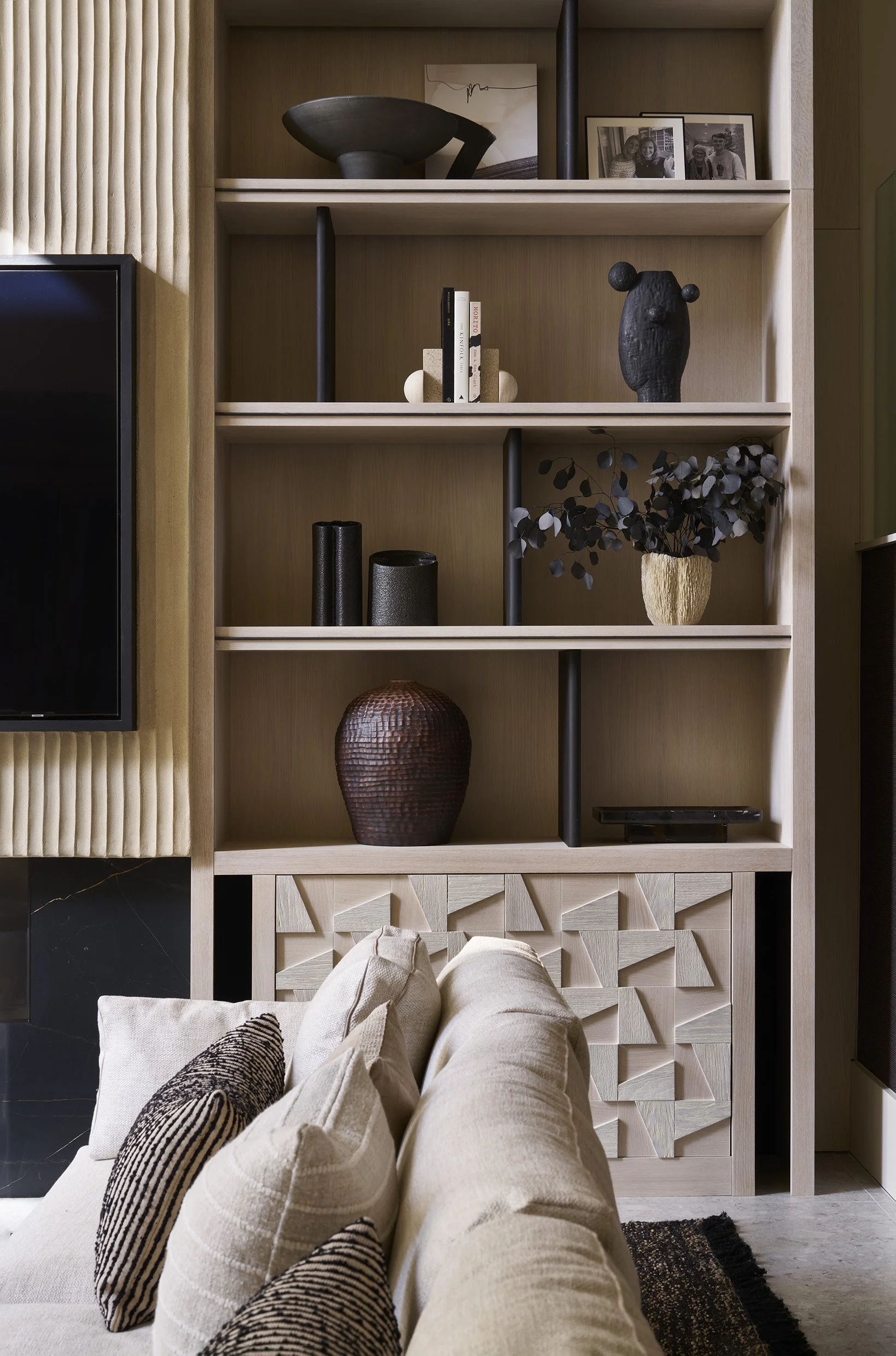 Close-up of a modern beige sofa with patterned throw pillows in front of a wooden shelf that displays black, white, and earthy-toned decorative objects, a framed photo, and books at the back of a contemporary living room.