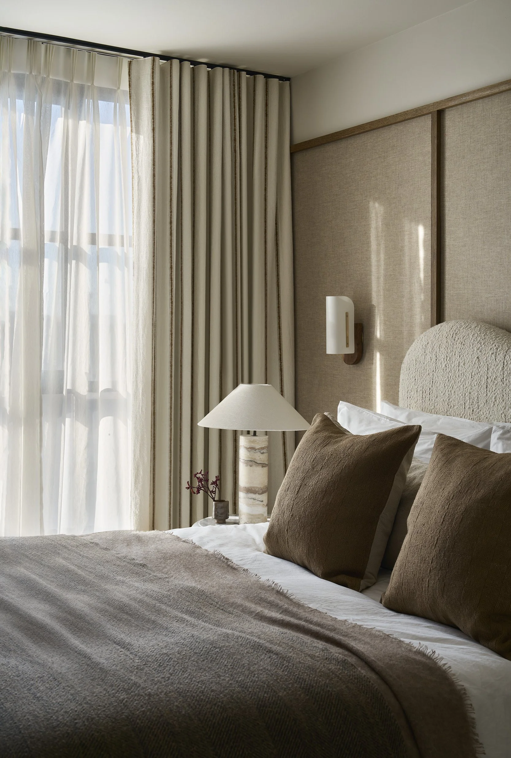 A neatly made bed with white sheets, beige headboard, and brown pillows in a hotel room with large window and cream-colored curtains.