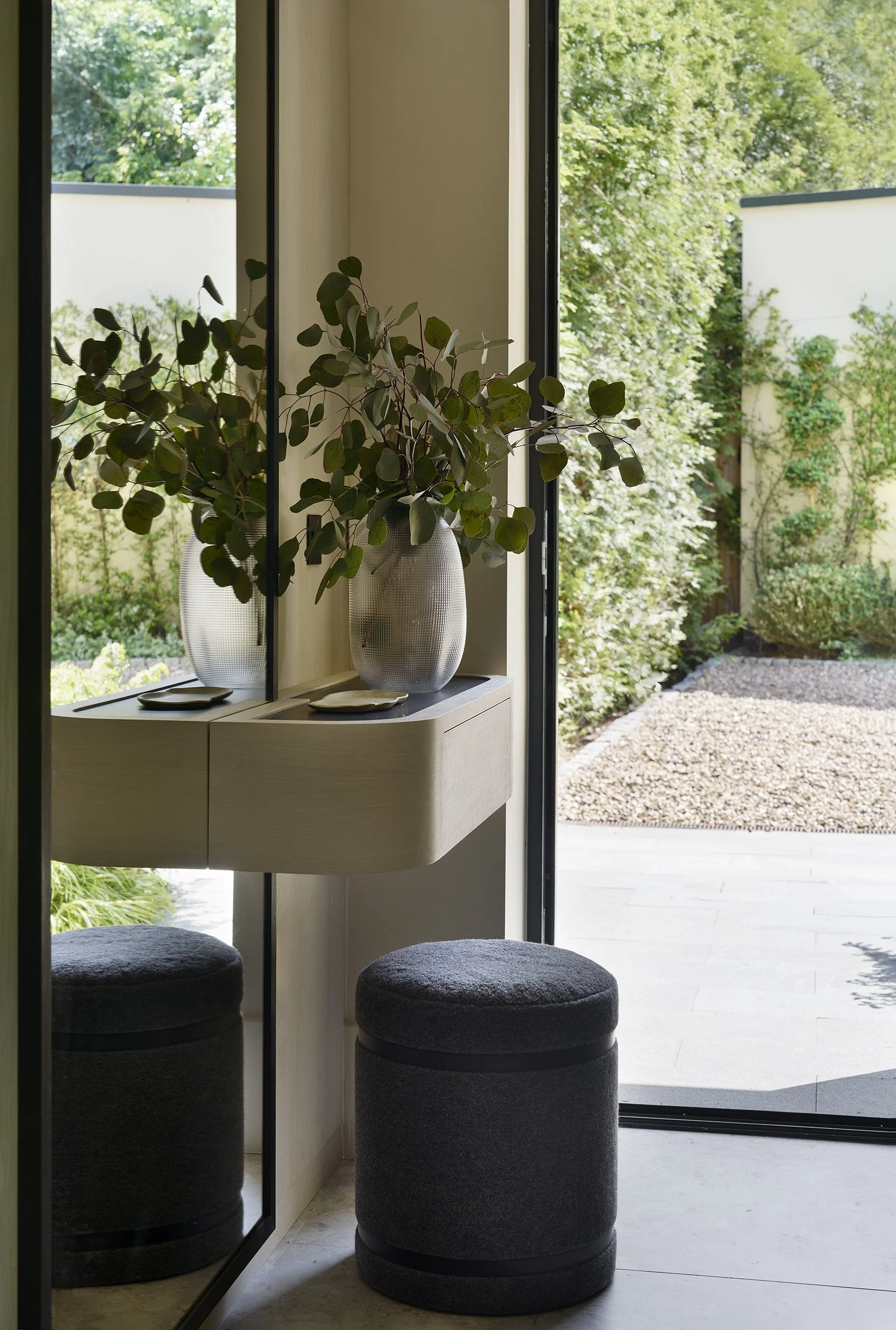 Interior view of a room with a small light-colored wooden table, a large potted eucalyptus plant, two black stools, and a sliding glass door opening to a garden with trees and gravel ground.