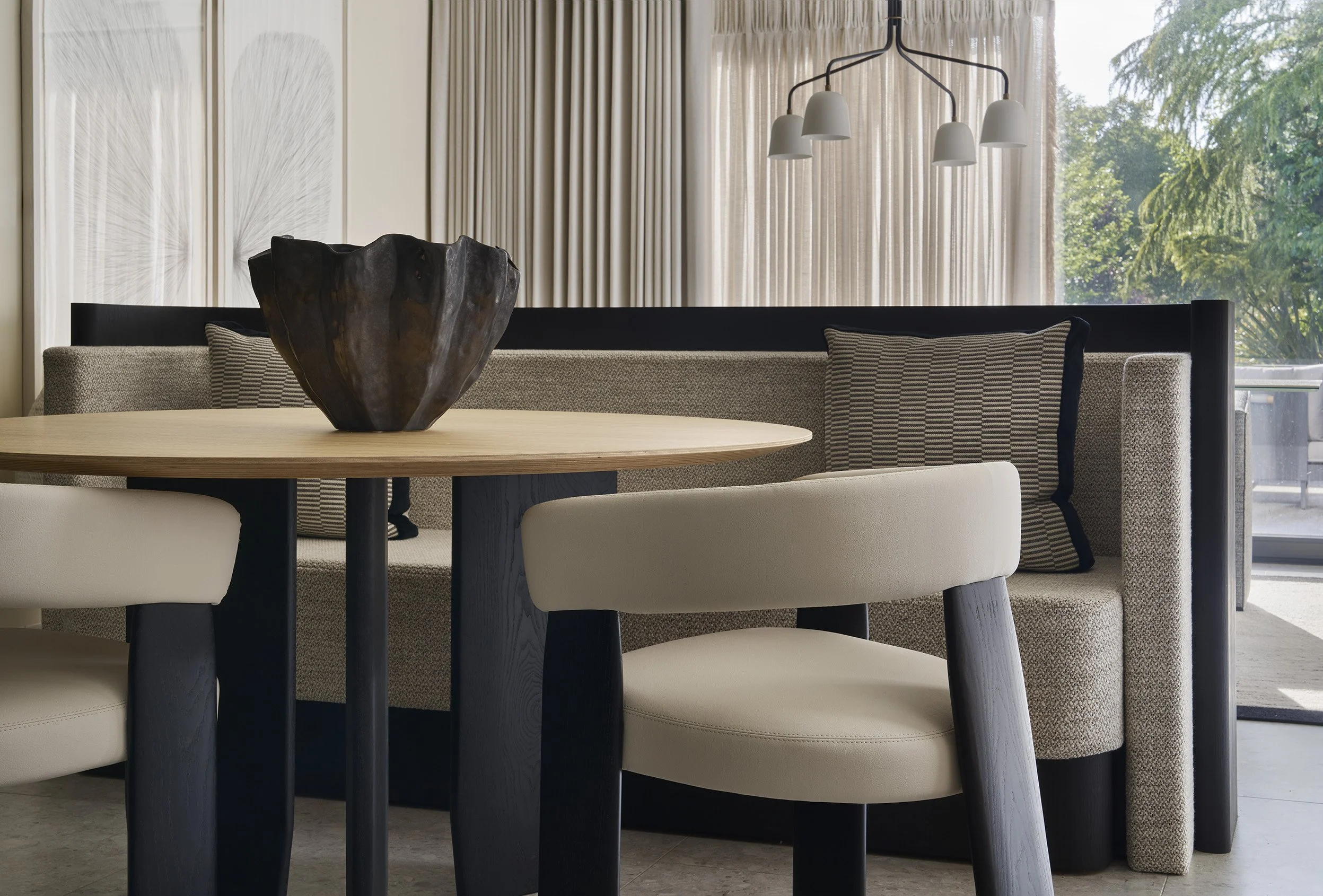 Modern dining area with a round wooden table, cream upholstered chairs, a built-in corner bench with pillows, a dark decorative bowl on the table, beige curtains, and large windows showing greenery outside.