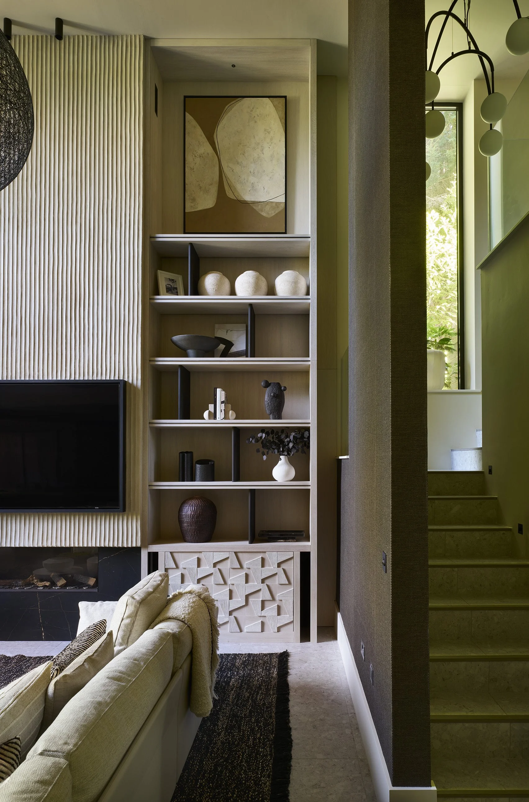 Interior of a modern living room with a built-in wooden shelving unit decorated with vases, art, and sculptures, adjacent to stairs and a window with sunlight.