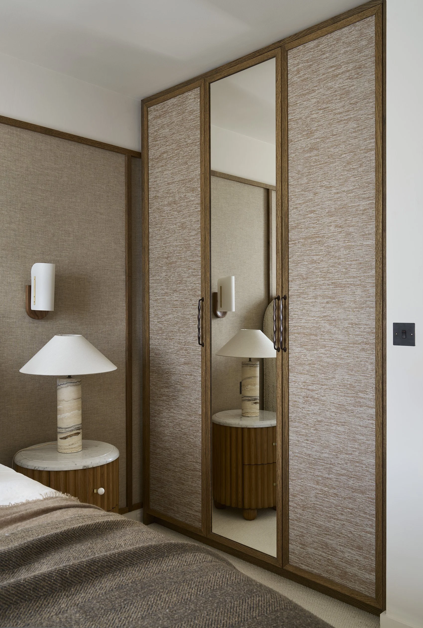 A bedroom corner featuring a brown and beige bedspread, two nightstands with marble bases, and a wardrobe with mirrored doors and wood trim.