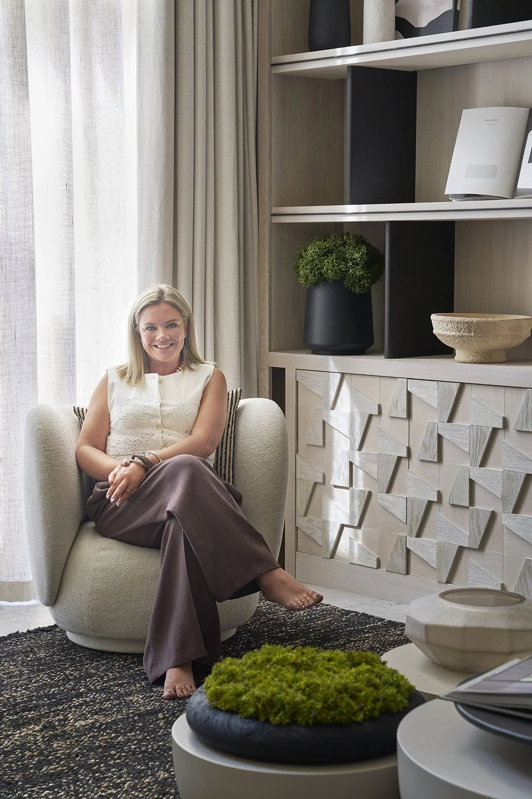 A woman sitting on a beige armchair in a modern living room with a wooden shelf and decor items, smiling at the camera.