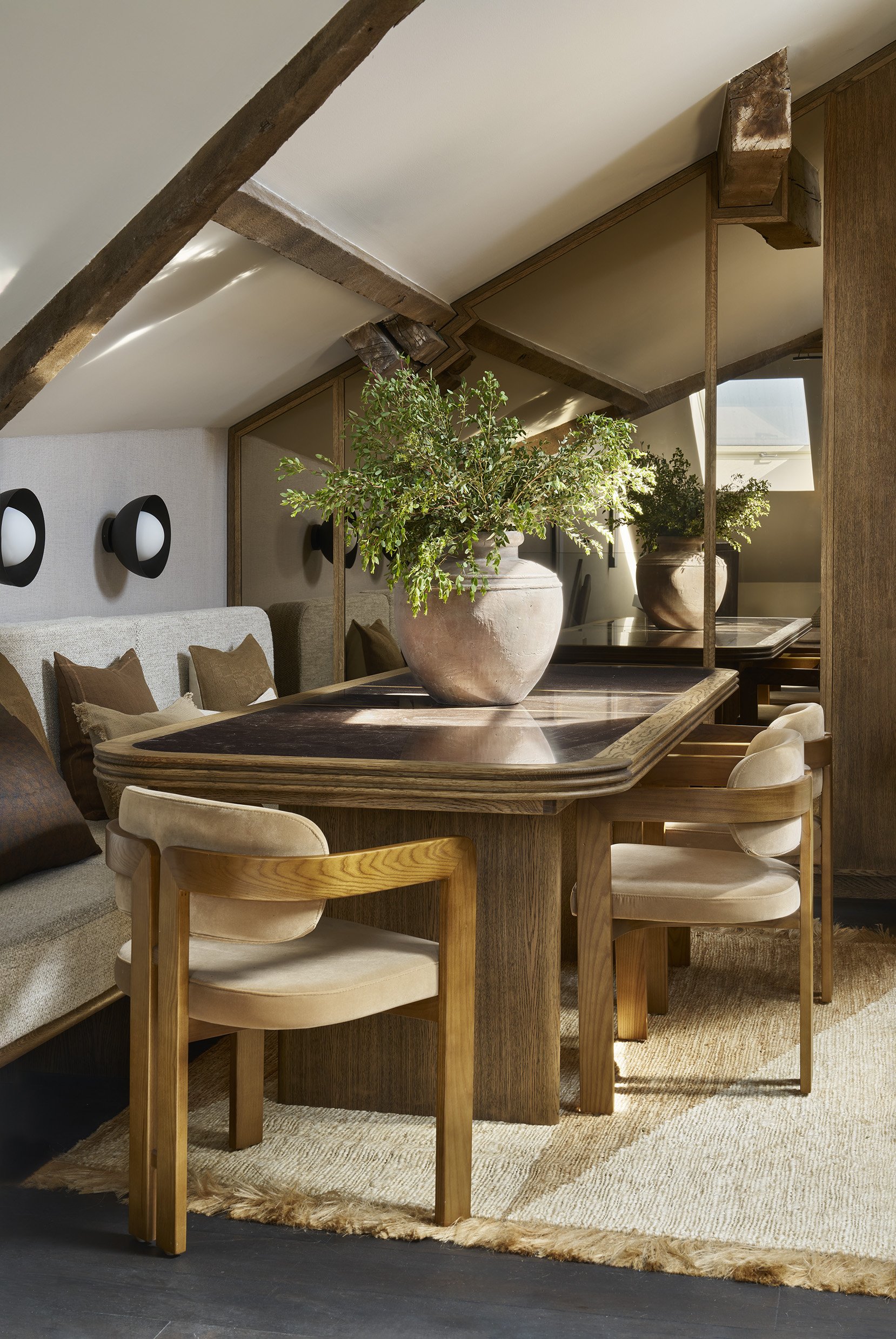 A cozy dining area with a wooden table, beige upholstered chairs, potted green plants, and wall-mounted lights under a sloped ceiling with wooden beams.