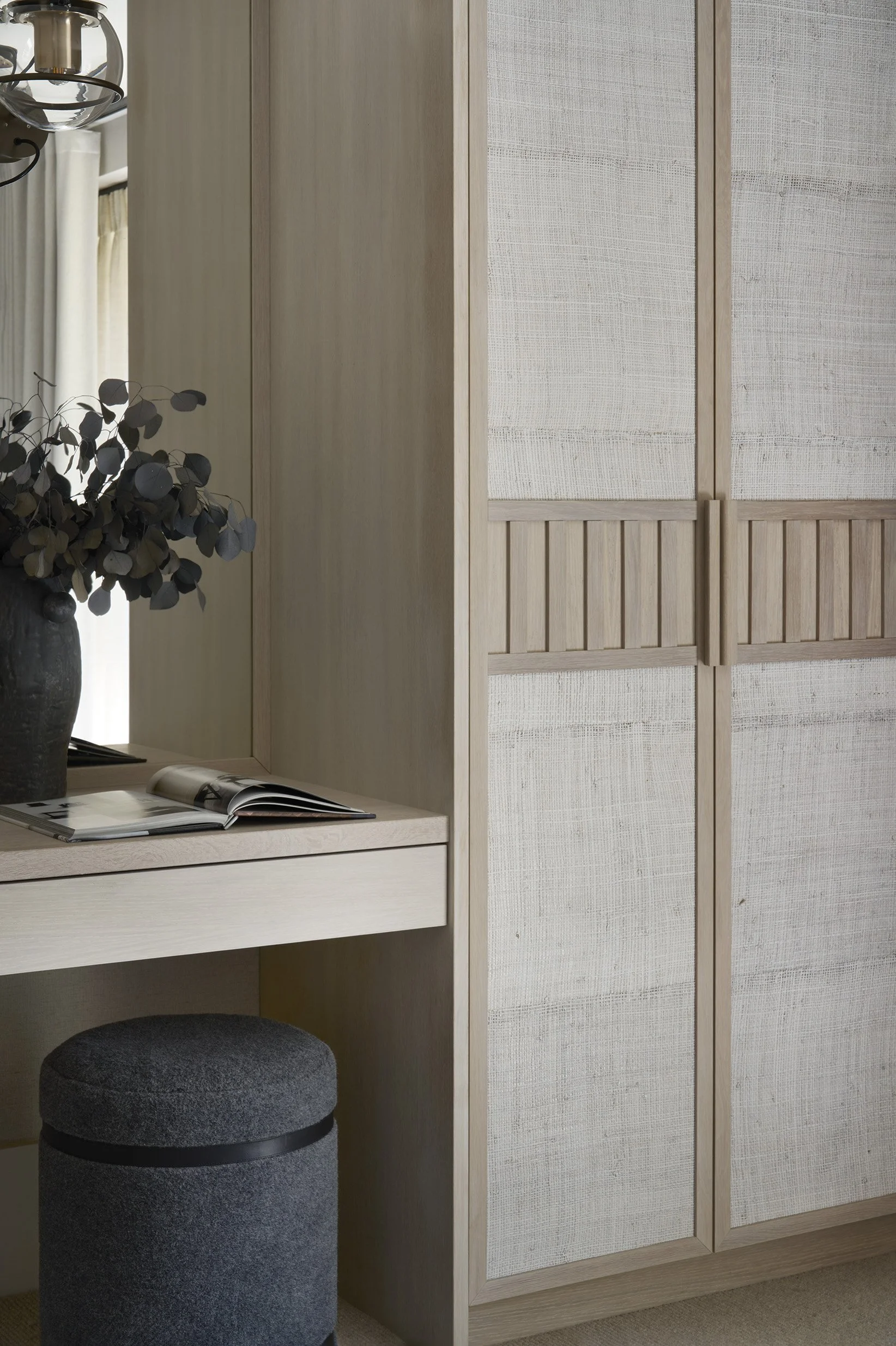 Wooden closet with sliding doors and a desk with an open magazine and a black vase with eucalyptus leaves. A gray upholstered ottoman is beneath the desk.