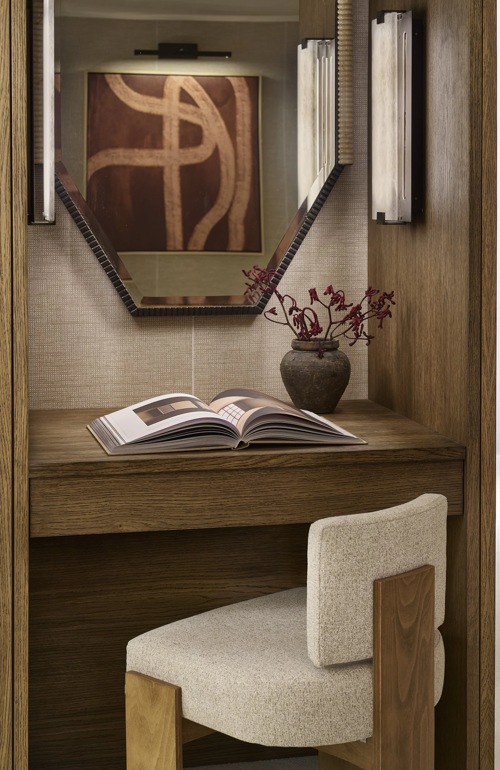 A small wooden desk with an open magazine, a brown vase with red decorative twigs, and a mirror mounted on the wall above. A beige upholstered chair is placed in front of the desk, and there is a piece of artwork visible in the mirror's reflection.