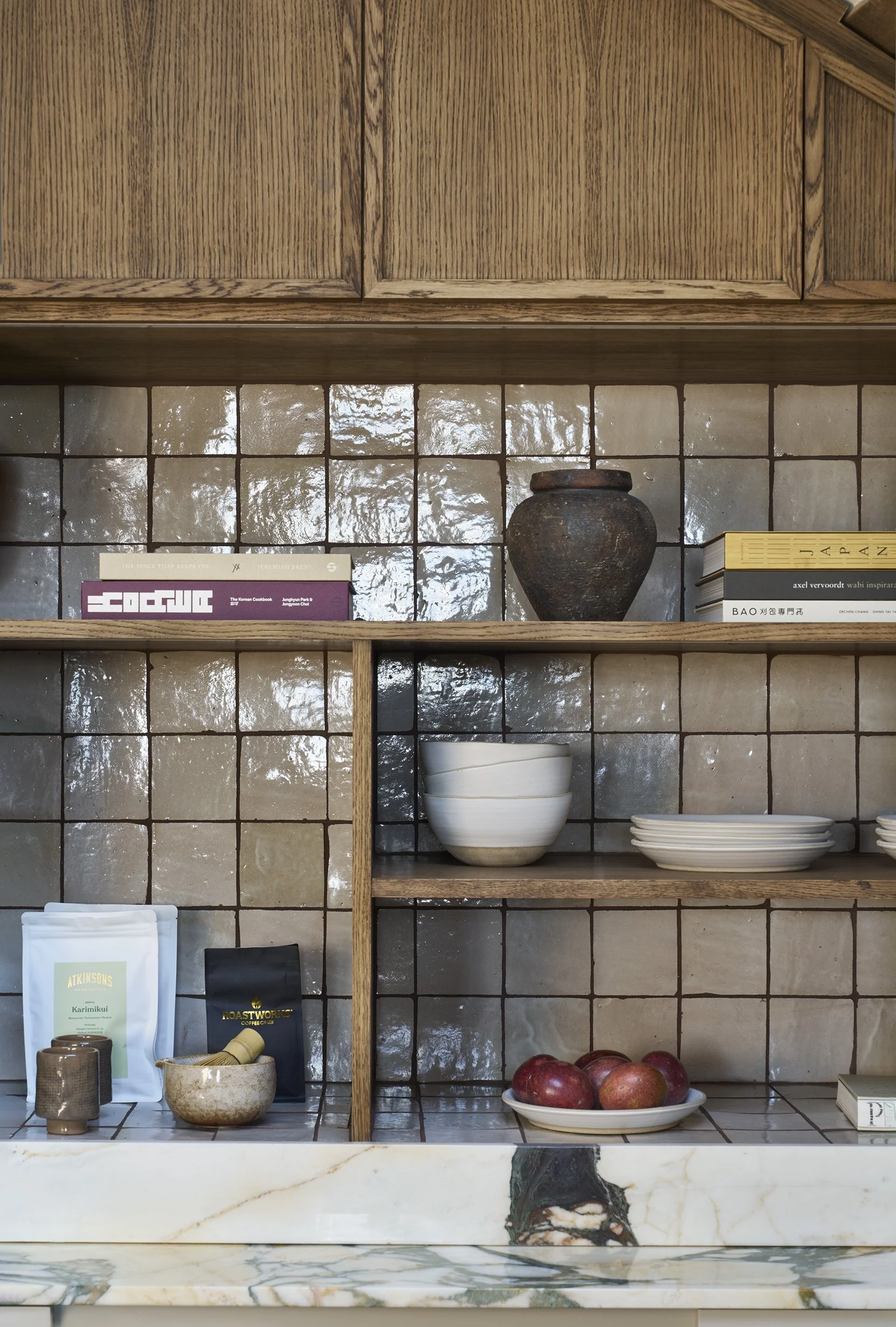 Kitchen shelf with books, a ceramic vase, bowls, plates, apples, and coffee bags against a tiled backsplash.