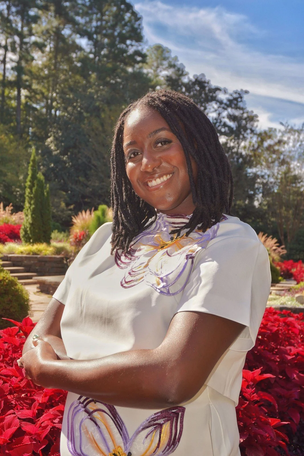Raven Ransom, trauma therapist, and a bright smile standing outdoors in a garden with red flowers and lush green trees, under a partly cloudy sky.