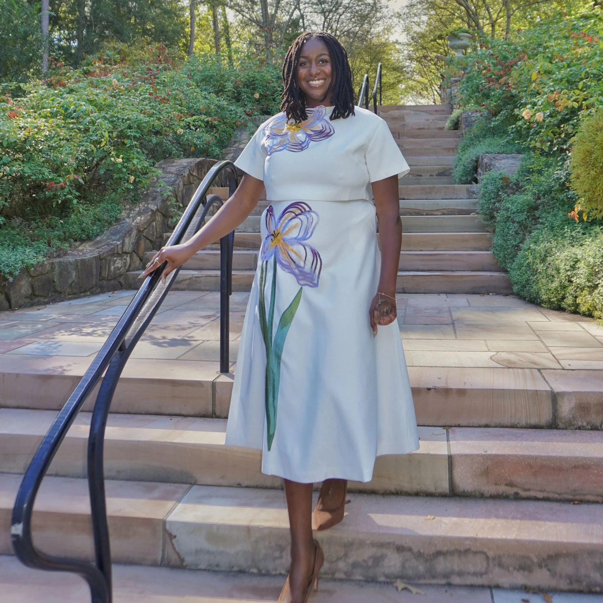 Raven Ransom, trauma therapist, standing confidently on outdoor wooden stairs in white dress with floral design, surrounded by lush greenery