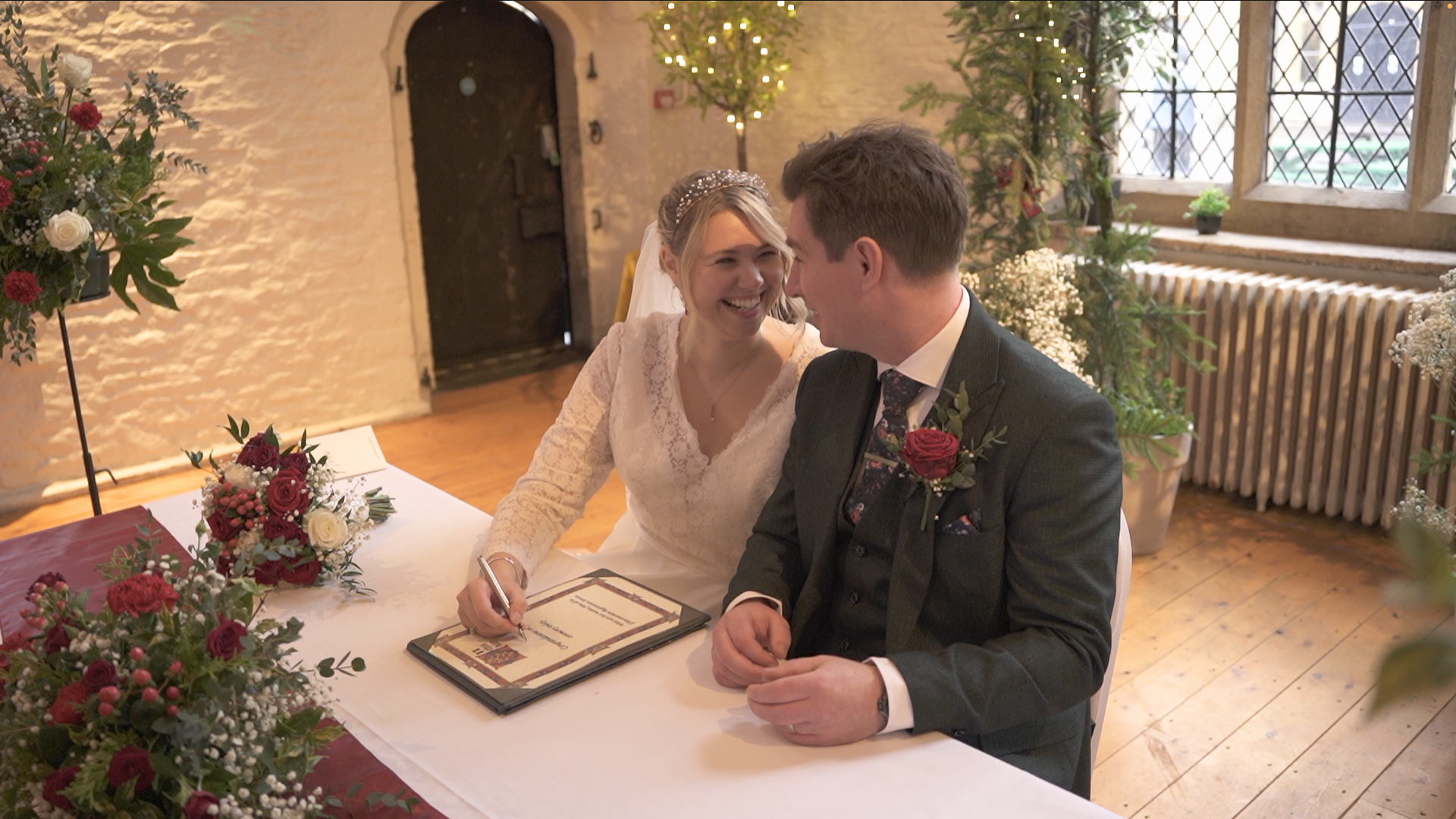 A bride and groom sitting at a wedding table, smiling and leaning close to each other, with the bride signing a document.