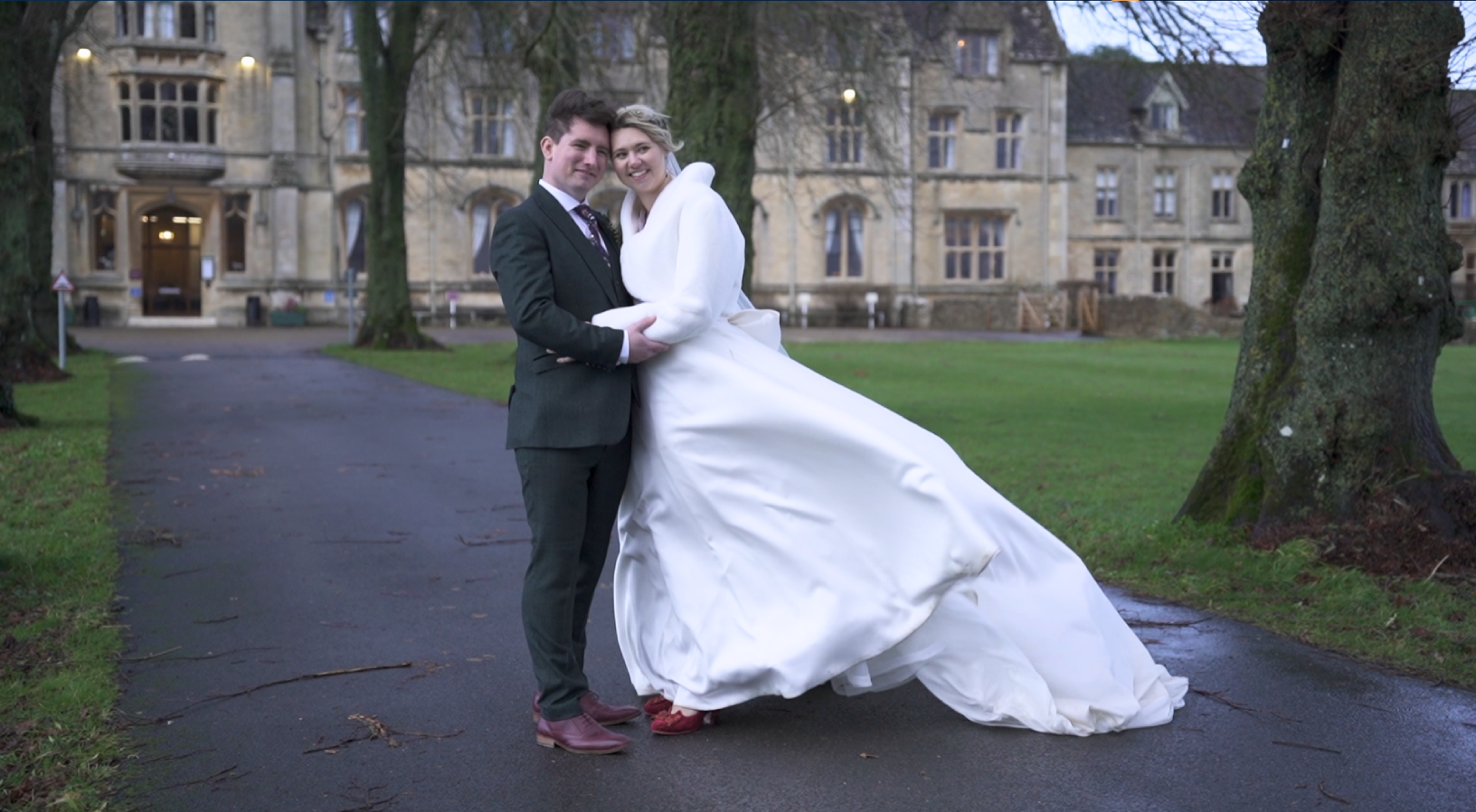 Bride and groom holding each other on a paved path in front of a large historic building, with trees on either side.