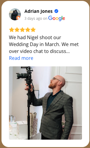 A man in a gray plaid suit with glasses and a beard is filming with a professional camera mounted on a tripod in a white room.