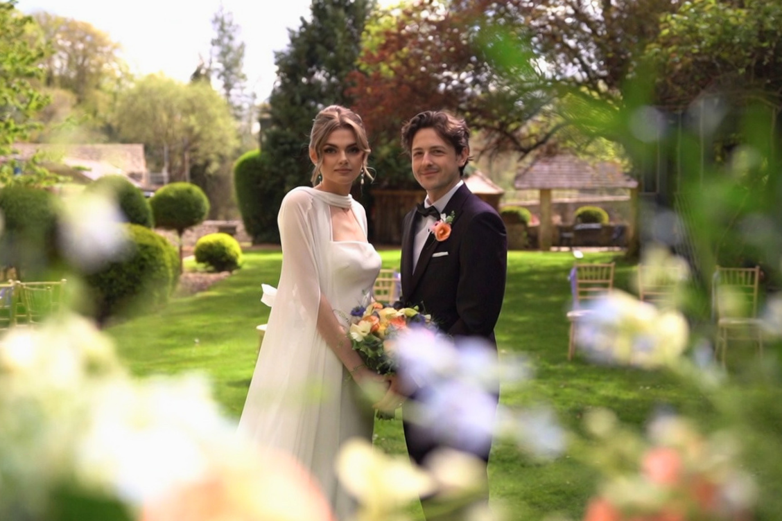 A bride and groom standing outdoors during their wedding, surrounded by greenery and trees. The bride wears a white gown and holds a bouquet of flowers, while the groom wears a black tuxedo with a pink rose boutonniere.