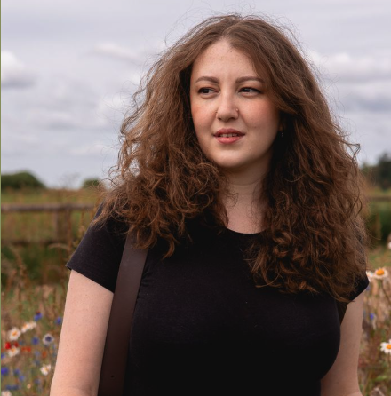 Young woman with curly hair standing outdoors in a field of flowers with cloudy sky.